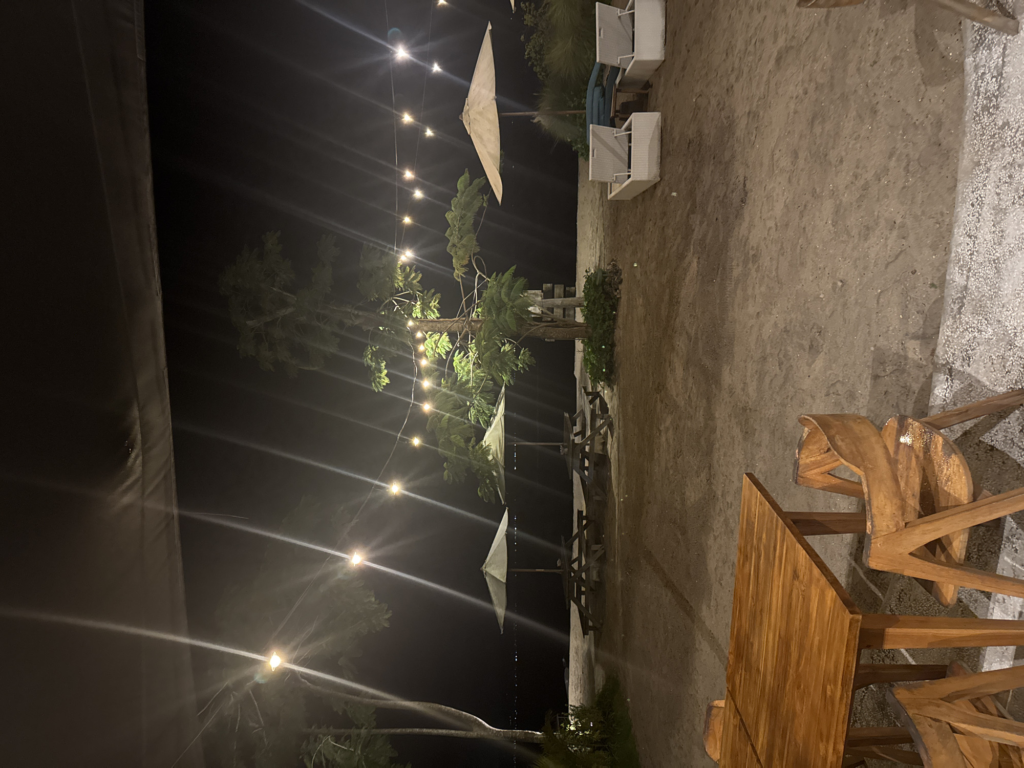 The photograph captures an outdoor dining area at night, likely located in Nusa Penida, Indonesia. Strings of lights are suspended overhead, illuminating the scene with a warm glow. Wooden tables and chairs are arranged on a sandy surface. Beach loungers can be seen in the background. The scene appears to be near a shoreline, with trees and vegetation framing the background. The night sky is dark, creating a contrast with the illuminated dining area. There are no people visible in the photograph.