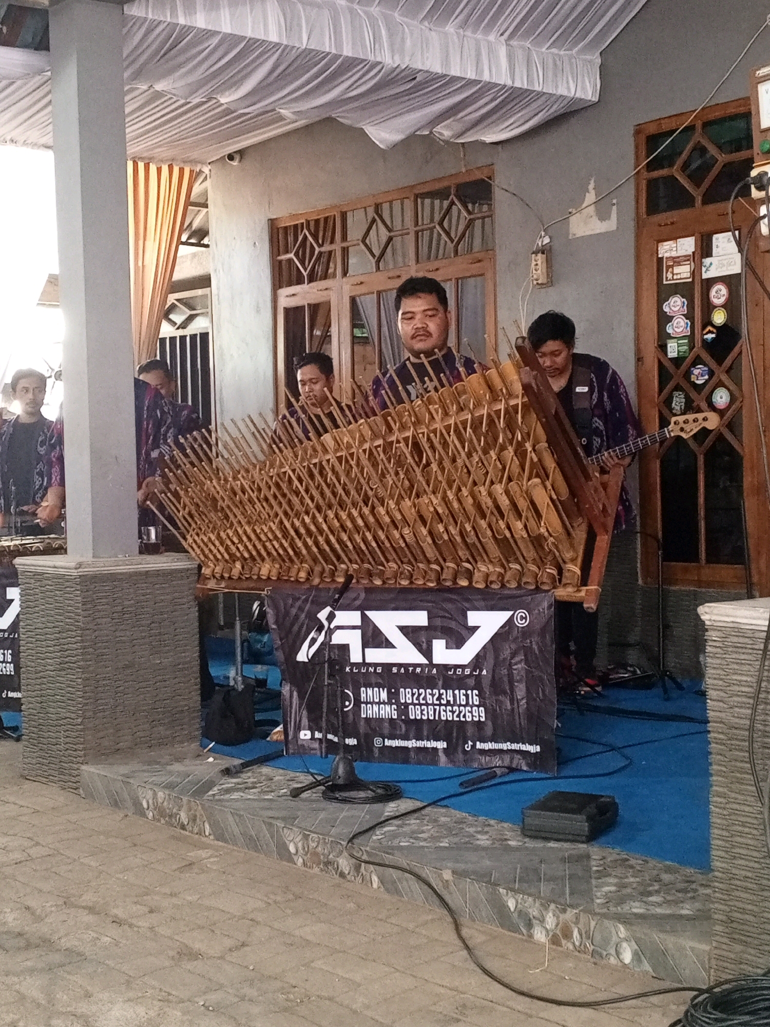 The image depicts a musical performance taking place outdoors, likely in front of a building with a covered patio. The setting appears to be Sedayu, Indonesia, as indicated by the text on a banner.
In the foreground, a group of men are playing traditional Indonesian musical instruments, likely angklung, which are bamboo rattles. The instruments are arranged in a large, tiered structure made of wood and bamboo. Several individuals are visible playing these instruments. One man, positioned centrally, is wearing a dark patterned shirt and appears to be the primary musician. To his right, another man in a similar shirt is playing a bass guitar. On the left side of the image, a person in a dark shirt is partially visible, seemingly also involved in the performance.
A black banner with white text and a logo is displayed prominently in front of the musical instruments. The text on the banner includes "KLUNG SATRIA JOGJA" and contact information. The ground beneath the performers is covered in blue carpeting, with the immediate area around the banner and instruments being a raised tiled platform. Various cables and a microphone stand are visible on the ground.
The background shows the exterior of a building with wooden doors and windows. A white canopy or tent is set up overhead, providing shade. There are also some decorative elements on the wall and hanging from the ceiling. The overall scene suggests a cultural event or a performance at a local venue.