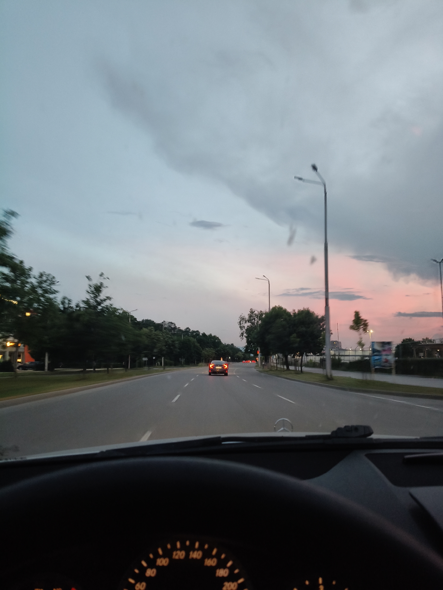 The image captures a view from inside a car, looking forward along a road at dusk. The sky is filled with clouds, transitioning from a pale blue to hues of pink and gray. Streetlights are visible along the roadside, as are trees lining the street. A few cars are present on the road, including a red car ahead and a dark car in the distance. The dashboard and steering wheel of the car from which the photo is taken are partially visible in the foreground, along with a speedometer indicating a speed between 60 and 80. The scene suggests a suburban or city environment during the evening commute.