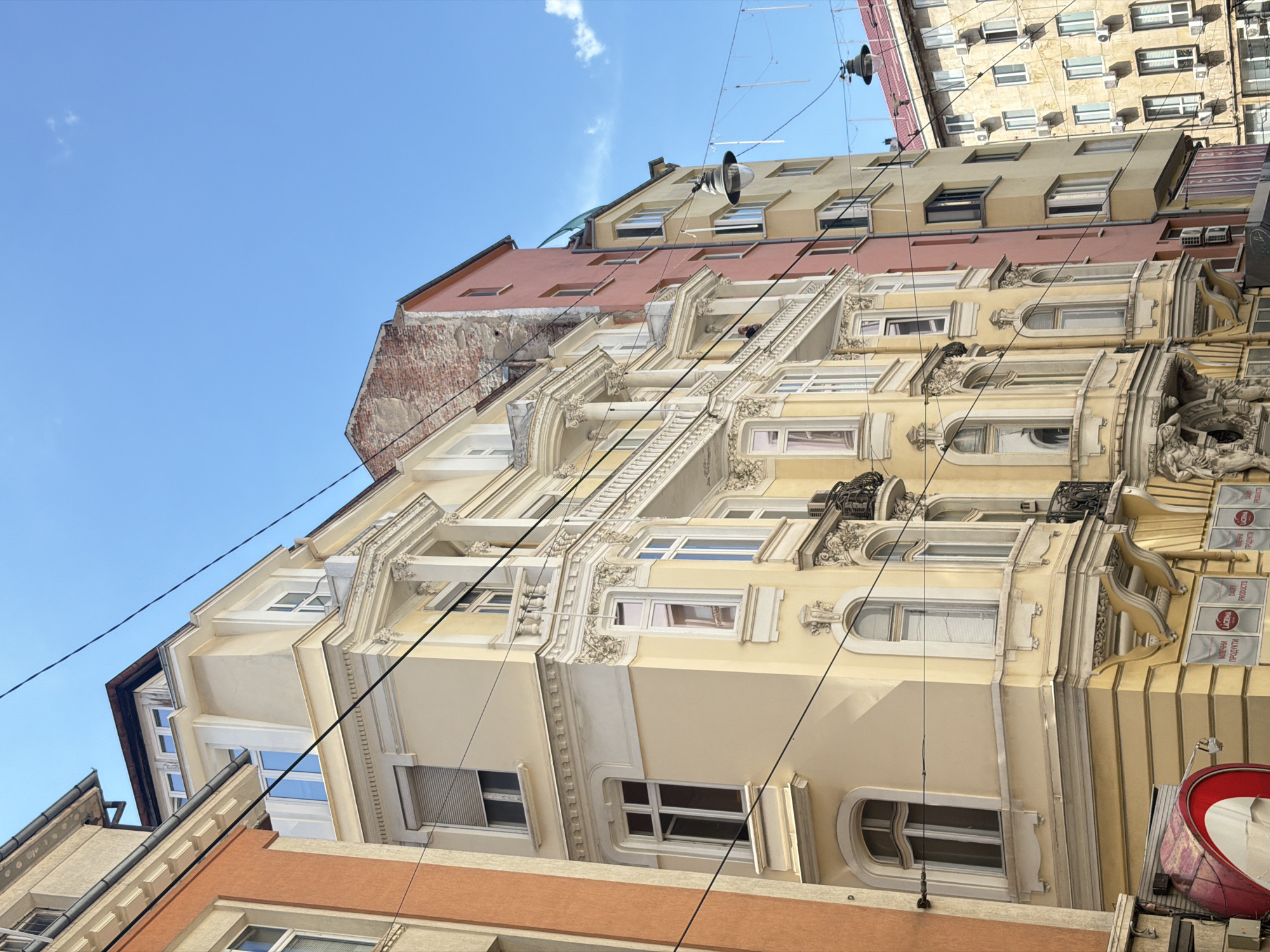 The image captures an upward-angled view of several multi-story buildings against a clear, bright blue sky with scattered white clouds, characteristic of an urban setting in an older European city.
The central and most prominent building is light yellow, showcasing ornate architectural details including decorative molding, balustrades, and varied window styles. It features arched and rectangular windows, some with small balconies adorned with wrought iron railings, and several visible air conditioning units. Below this, part of an orange/terracotta-colored building is visible. Behind the yellow building, a reddish-brown structure with exposed brickwork and simpler rectangular windows can be seen. Further in the background, another lighter-toned building with numerous, more modern windows and external blinds extends upwards.
Numerous black power lines or communication cables crisscross the frame, running between the buildings. A large, dark, conical loudspeaker hangs from one of these wires, pointing downwards. On the lower right, a large red circular sign is partially visible.
Visible text on the central yellow building includes signs for "LACTIMA" and "МЛЕЧНИ ПРОДУКТИ," which translates to "Dairy Products" in Bulgarian, suggesting a commercial establishment. No people are clearly visible. The bright lighting indicates a sunny daytime scene. The architecture suggests a historical European urban environment, consistent with the location context of Sofia, Bulgaria.