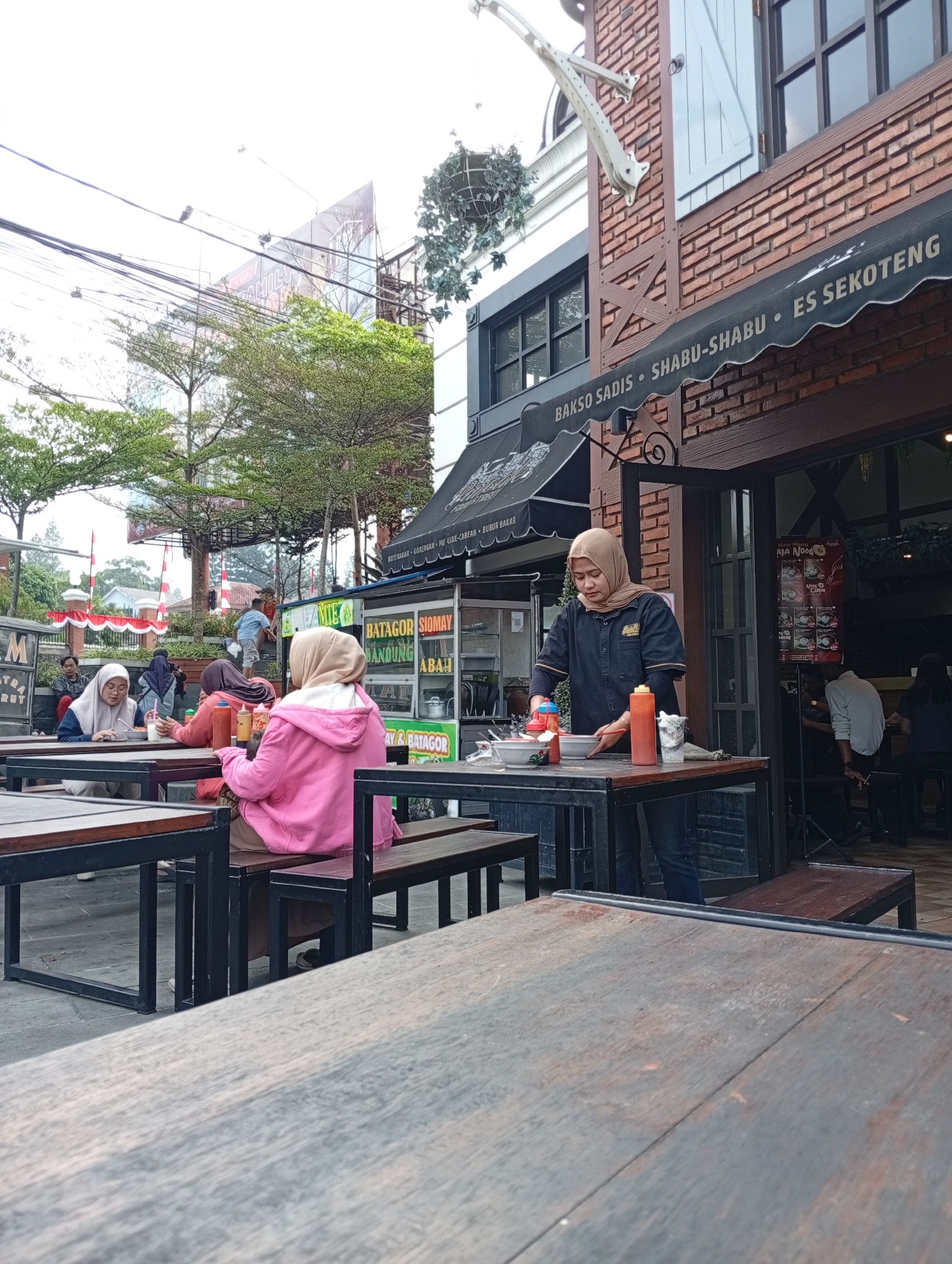 The image captures an outdoor dining area in front of a brick building, likely a restaurant or food stall, in Cimacan, Indonesia. A black awning above the entrance displays text including "BAKSO SADIS," "SHABU-SHABU," and "ES SEKOTENG," indicating the types of food and drinks offered. A woman wearing a beige hijab and a dark uniform is preparing food at a table, with bowls, bottles of condiments, and a glass of liquid visible. Several people are seated at tables in the foreground and mid-ground, some eating and others conversing. A woman in a bright pink hoodie is visible from the back, facing away from the camera. Behind the dining area, a street scene unfolds with trees, utility poles and wires, a billboard, and buildings in the distance. Red and white flags are also present, suggesting a local event or decoration. The overall scene depicts a casual dining environment in a moderately populated urban or semi-urban setting.
