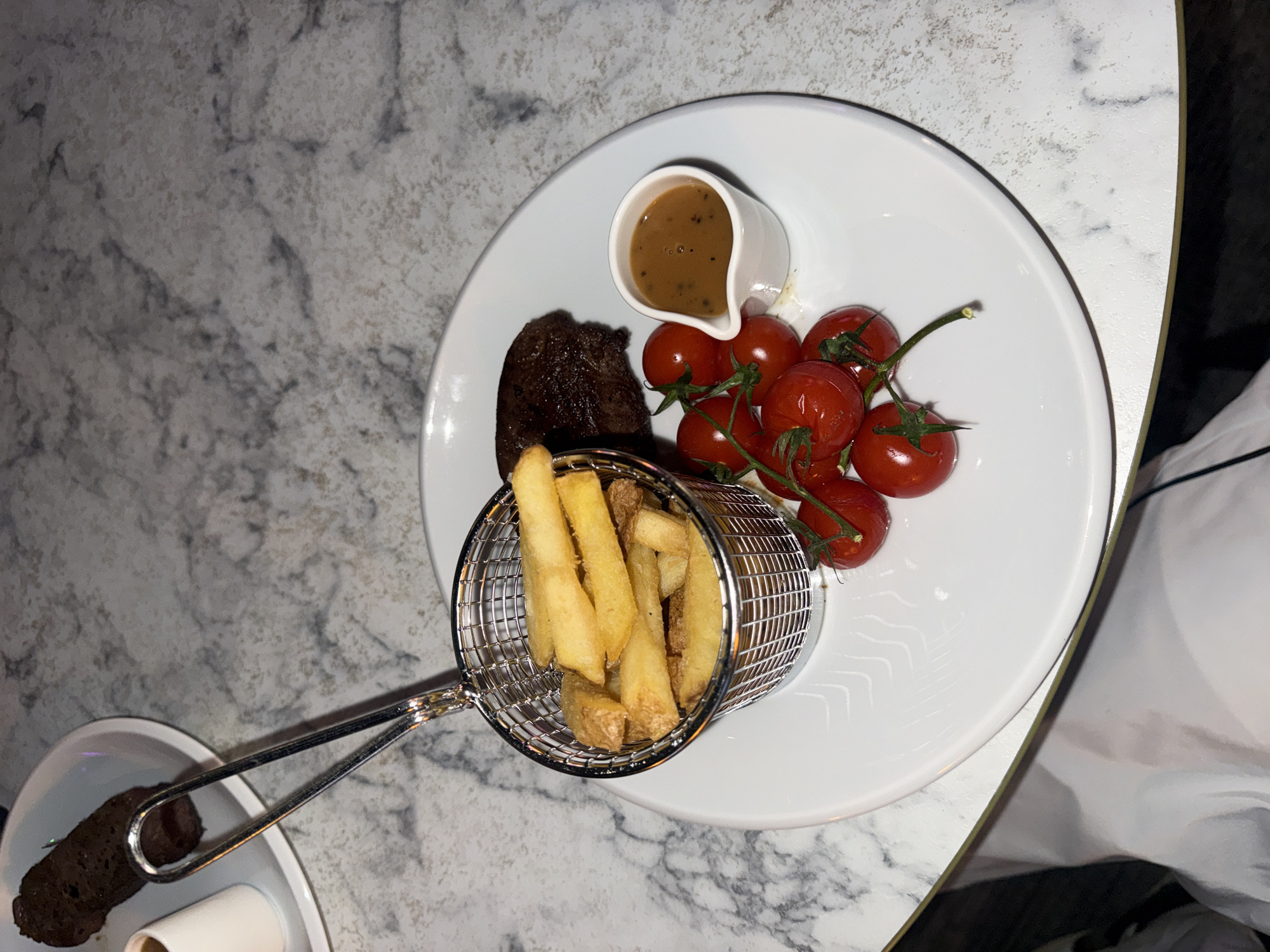 An overhead shot shows a white plate with a steak, fries in a mini metal basket, cherry tomatoes on the vine, and a small pitcher of sauce. The food is on a white plate with a subtle etched pattern. The plate rests on a marble countertop. In the background, partially visible on another white plate, is a piece of dark meat. The scene is dimly lit.