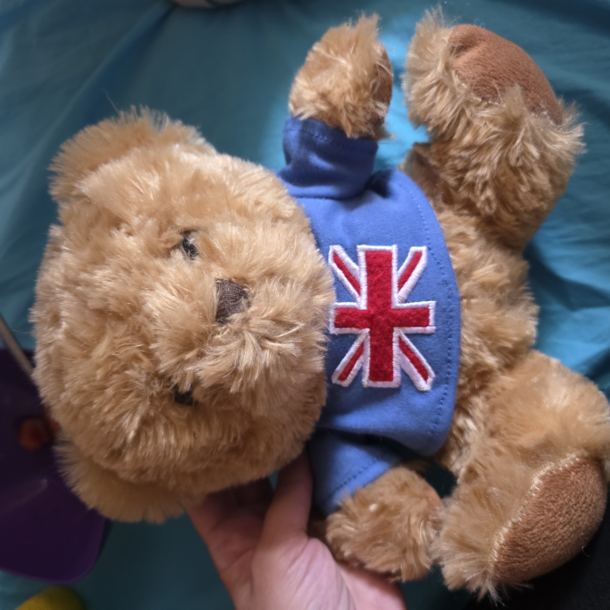 A close-up, eye-level shot shows a light brown, plush teddy bear sitting upright. The bear is wearing a bright blue t-shirt with an embroidered Union Jack flag on the front. Its fur is soft and slightly tousled. A hand, with visible fingers and fingernails, is gently holding the bear from the left side, supporting its arm. The bear is positioned on a turquoise fabric surface, possibly a tablecloth or bedspread, which has some creases and folds. In the upper left corner, out of focus, is a purple bowl containing a few small, reddish-orange objects. The background is primarily the turquoise fabric. The scene is well-lit, with a slight shadow cast on the bear's face from the viewer's perspective. This image was taken in Smolevo, Bulgaria.