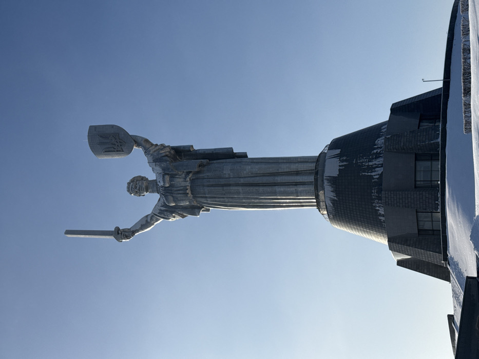 A large, silver-grey metallic statue, identified as the Motherland Monument (also known as Mother Ukraine), stands prominently against a clear, bright blue sky. The monument is located in Kyiv, Ukraine, specifically within the National Museum of the History of Ukraine in the Second World War complex. The colossal female figure faces forward, with her right arm raised, holding an upright, elongated sword. Her left arm is also raised, supporting a shield embossed with the Ukrainian national emblem, the Tryzub (trident).
The statue rests on a multi-tiered, grey architectural pedestal. Traces of white snow or ice are visible on the ledges and surfaces of the pedestal's upper sections, indicating cold weather conditions. The ground surrounding the base of the monument is covered in a layer of uniform white snow. In the lower right foreground, a patch of dark, low-lying shrubbery is partially visible, contrasting with the snow. A dark, slender pole, possibly a light fixture, extends vertically from the snow-covered ground near the base of the monument on the right side. No human figures are present in the scene. The overall lighting suggests a clear, sunny winter day.