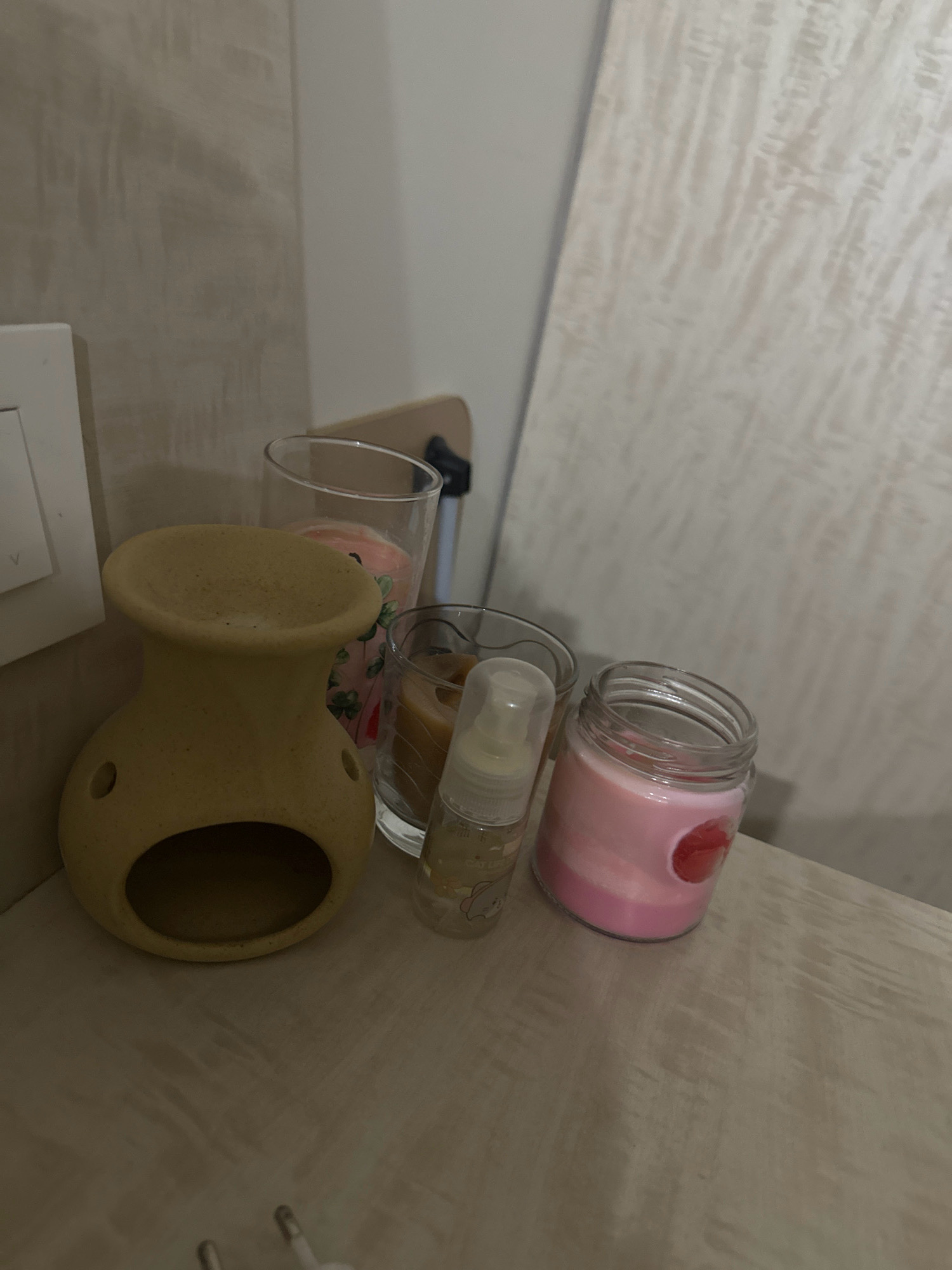 A dimly lit, close-up indoor shot shows a collection of decorative items on a wooden surface. The objects include a beige ceramic oil burner with a hollow interior and two circular openings on its side, a glass jar containing a pink candle with a red heart-shaped embellishment, a clear glass with a pink candle adorned with floral designs, and a small clear plastic bottle with a white pump dispenser and a yellow label. The background features a textured, light-colored wall and a portion of a light-colored paneled surface. The scene appears to be set on a shelf or tabletop in a domestic setting, possibly a bedroom or living area in Lalitpur, Nepal.