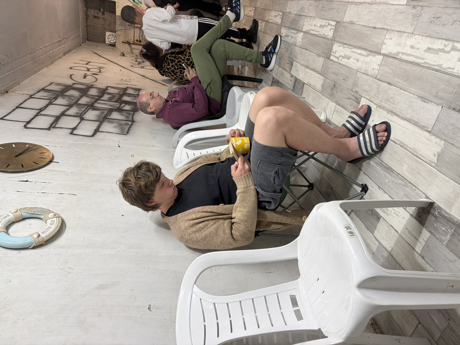 The image depicts an indoor waiting area with four individuals. The environment features a light grey concrete-like wall adorned with a pale blue and white lifebuoy labeled "HILTON HEAD" and a brass-colored clock. Graffiti, including a stylized brick pattern and the letters "G.B.H.", is present on the wall. The floor is covered with light-toned, wood-look plank flooring.
In the immediate foreground, a person with light brown hair, wearing a black t-shirt, light brown cardigan, dark grey cargo shorts, and blue-and-white striped slide sandals, sits on a black-framed folding chair. They hold a yellow mug with a white spoon, directing their gaze downwards. An empty white plastic chair stands to their left.
In the midground, on the right side, an adult male with short grey hair and glasses, dressed in a maroon long-sleeved shirt and olive-green pants, sits on a white plastic chair, focused on a smartphone. His left leg is crossed over his right. Behind him, an adult female wearing a leopard-print top is partially visible, also appearing to interact with a smartphone. In the background, a younger male in a white long-sleeved shirt with black stripes on the sleeves is standing. No direct interactions among the individuals are depicted. The precise location within Tel-Aviv, Israel, is not identifiable from the visual information provided.