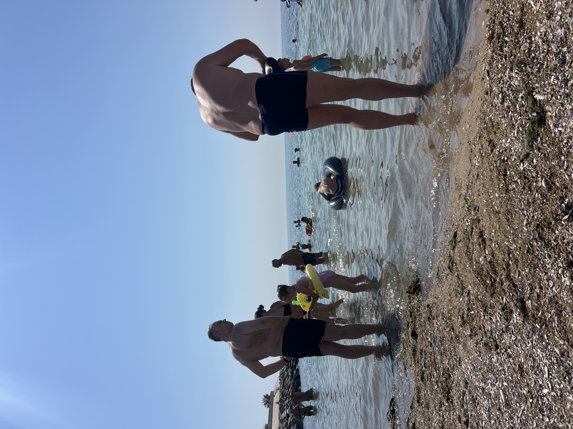 The image captures a sunny day at the beach in Fontanka, Ukraine, with a clear blue sky and calm, shallow water. Several people are enjoying the water and shoreline. In the foreground, a man in black swim trunks stands with his back to the camera, his head and upper torso slightly blurred, suggesting a dynamic pose or movement. The water laps at his feet, and the sandy, shell-strewn shore is visible to his right.
Further into the water, a man is partially submerged, holding onto a black inner tube. In the middle ground, a man in blue swim trunks stands in the water, looking up, while a child in blue shorts stands beside him. A group of people, including a woman in a black swimsuit and a child in a pink swimsuit with a yellow inflatable toy, are also in the shallow water.
In the background, more people are scattered throughout the water, some swimming and others standing. The scene suggests a relaxed and casual atmosphere, typical of a summer day at the beach. The lighting indicates it is likely daytime, with the sun high in the sky. No text is visible in the image.