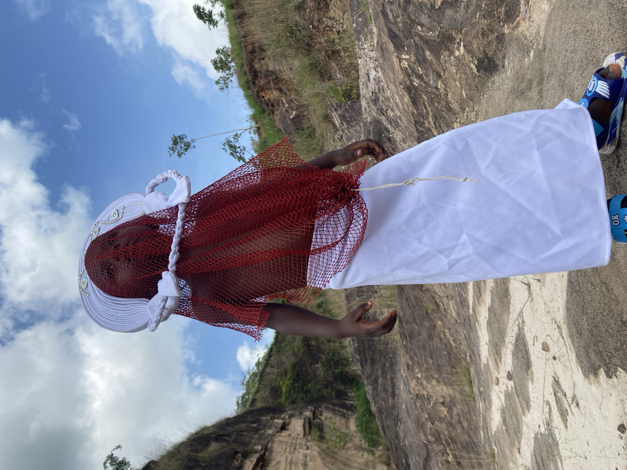 An image, originally rotated 90 degrees to the left, depicts a person standing outdoors against a dramatic rocky landscape in Achara, Nigeria. The individual, appearing to be a child or young person, is dressed in striking attire. They wear a wide-brimmed white hat or headpiece, possibly pleated, adorned with shiny embellishments on the brim and braided white accents. Their upper body is covered by a vibrant red, open-weave mesh garment, while a plain white, voluminous lower garment, possibly a skirt or wrap, is tied with a simple string around the waist. On their feet are blue and light blue patterned sandals, one of which visibly displays the text "OK" on its strap.
The person stands with both arms slightly bent at the elbows, forearms extended forward, and palms open and facing upwards, suggesting a pose that could be ceremonial, receptive, or performative.
The background features a steep, rugged rocky hillside on the left, covered with patches of grass and sparse green trees under a bright blue sky with scattered white clouds, indicating daytime. To the right of the person is a large, light-colored rock formation or cliff face with distinct vertical textures and streaks. The ground where the person stands appears to be a mix of sand and gravel.