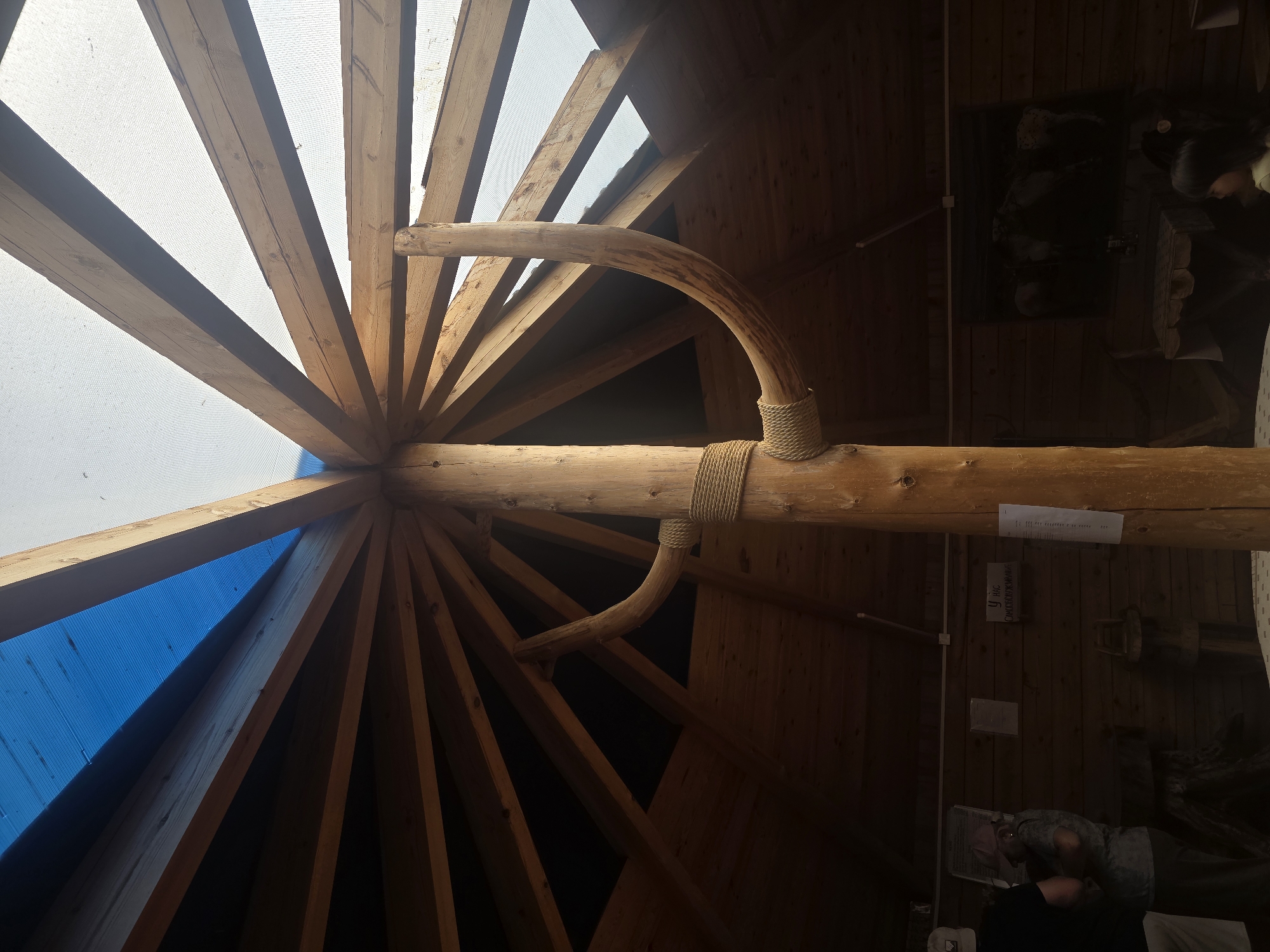In Maksimikha, Russia, this image captures the interior of a wooden structure, likely a gazebo or a covered outdoor seating area, characterized by a central, thick wooden post supporting a radial roof structure. The roof is comprised of numerous wooden beams extending outwards, with sections covered by both a translucent white material and a bright blue tarp. A curved wooden branch-like element is attached to the central post, secured by rope. The walls of the structure are made of horizontal wooden planks. In the background, on the wall, are several signs and a framed picture. To the left, two individuals, one wearing a cap and the other a pink hat, are partially visible, interacting near a wooden display. To the right, a young girl with dark hair is seated, looking to the side. Various wooden furniture and objects are present within the space, suggesting a functional or recreational area. The lighting is dim, with sunlight filtering through the translucent roof sections.