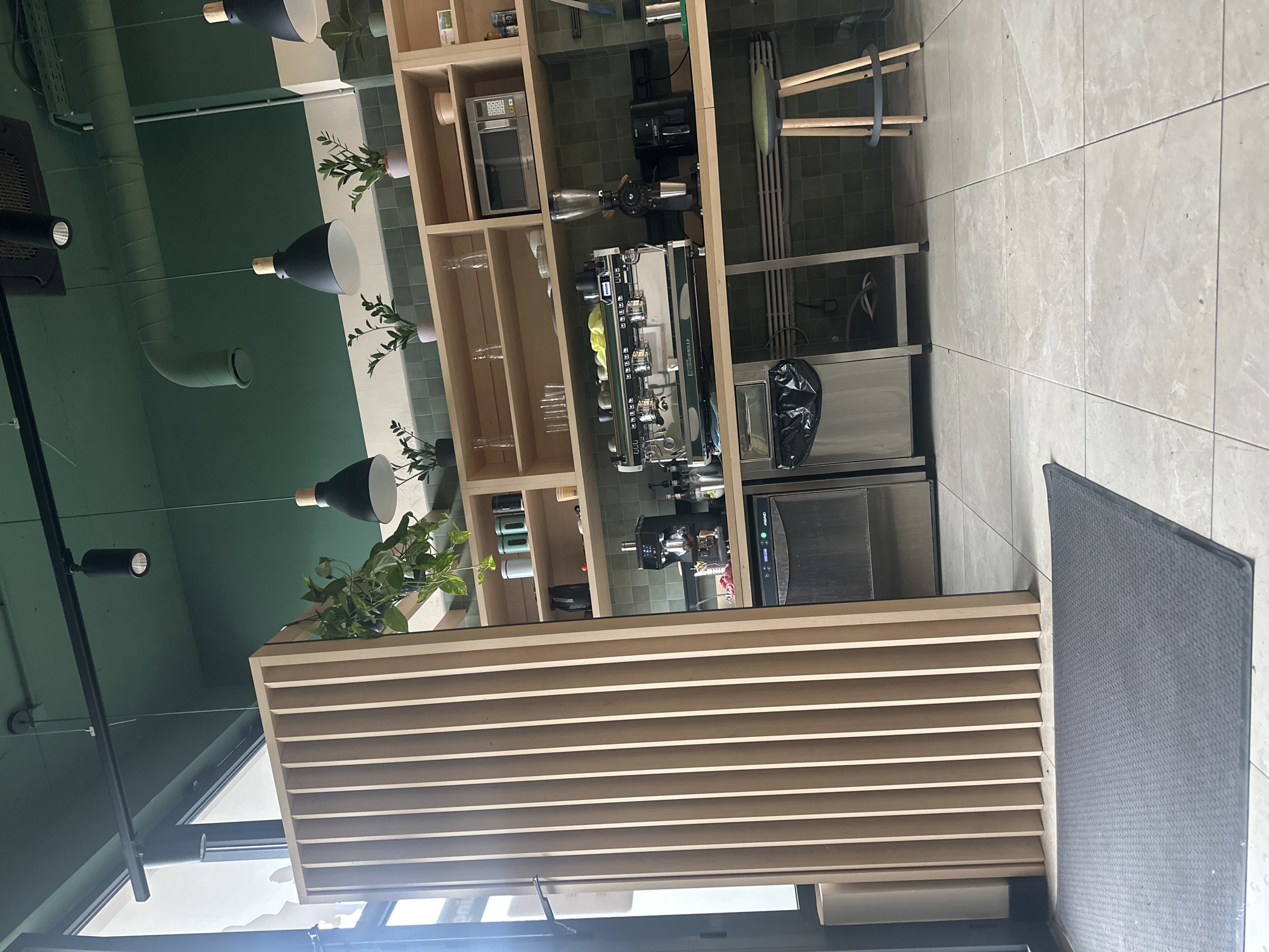 This image captures the interior of a modern cafe or bar, characterized by clean lines and functional design. The setting features a prominent coffee station against a wall painted in a dark teal or deep green hue.
In the background, a wooden shelving unit houses various items, including a professional-grade espresso machine (identifiable as "LA MARZOCCO"), a coffee grinder, and a microwave oven. Several potted green plants are placed on the shelves, adding natural accents, alongside neatly stacked glassware. Below the coffee equipment, stainless steel units, possibly refrigeration or storage, are visible, with a black trash bag placed beside one.
The ceiling, also painted green, features exposed ductwork and modern black pendant lights with wooden accents, as well as spotlights. Natural light appears to enter from a large window or glass partition on the left side of the frame.
In the foreground and midground, a slatted wooden counter or bar structure runs horizontally across the image. The floor is covered with large, light grey tiled squares. Partially visible on the right is a black, textured surface, possibly a tabletop or a seat. A stool with wooden legs and a green seat can be seen behind the coffee station.
No people are present, suggesting a quiet moment in the establishment. The lighting indicates it is daytime.