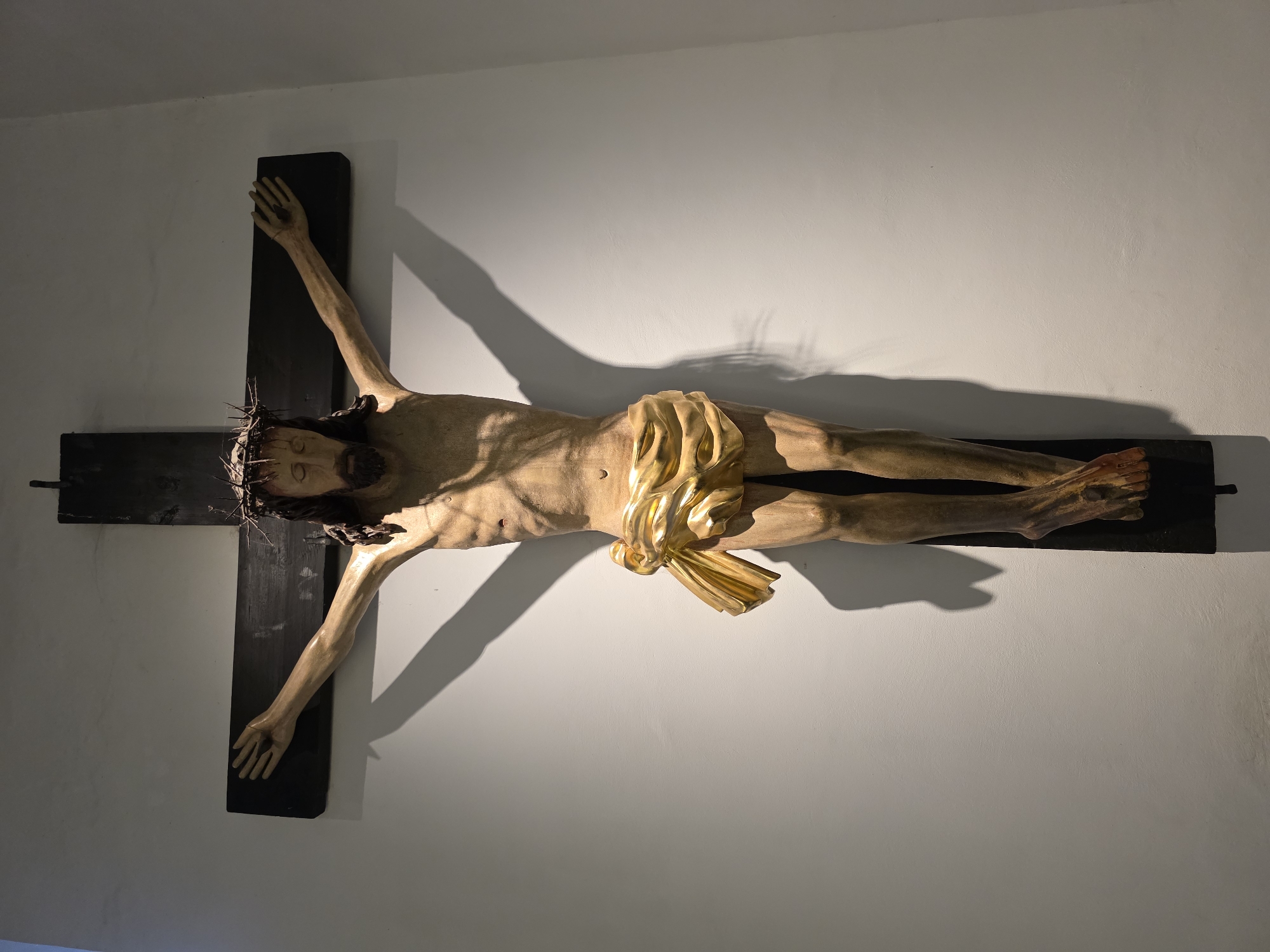 This is a full, head-on, medium shot of a wooden crucifix mounted on a white wall. The crucifix depicts Jesus Christ with a crown of thorns, a golden loincloth, and nails through his hands and feet. The lighting casts a prominent shadow of the crucifix on the wall to the right. The scene is devoid of people, action, or interaction. There is no discernible information within the image to indicate its location in Warsaw, Poland.