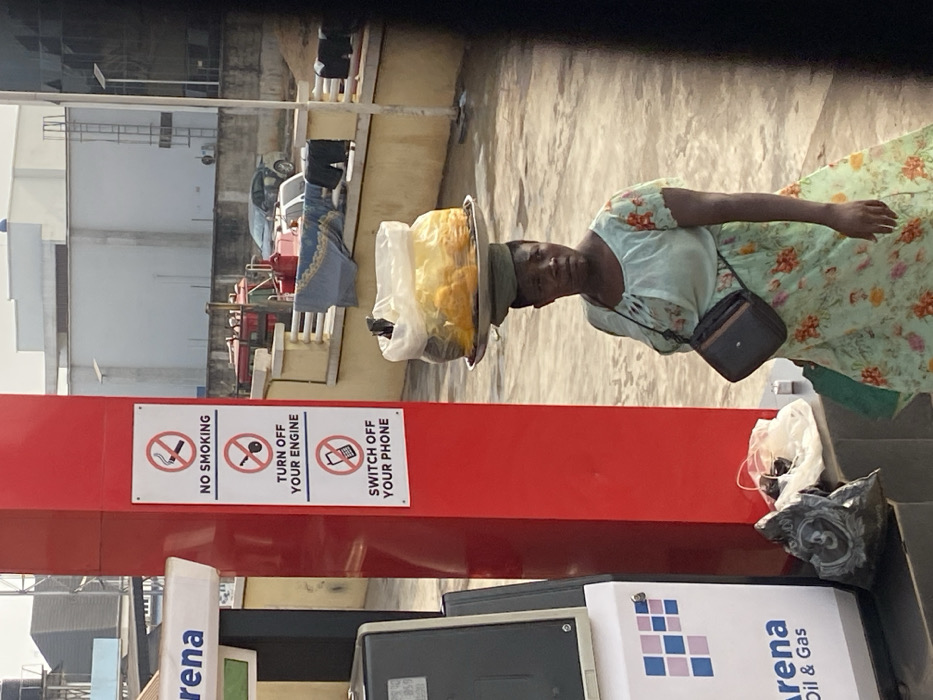 The image captures an outdoor scene in Issele Uku, Nigeria, under bright daylight. A dark-skinned woman is positioned in the midground, slightly to the right of the frame's center, facing partially towards the right. She wears a light green dress with a floral pattern of orange, green, and pink, and has a small black rectangular purse with a strap slung across her body. She balances a round, metallic tray on her head, which holds a large clear plastic bag filled with numerous yellow or orange spherical fruits, and a dark, unidentifiable object. Her head is covered by a green cloth.
To the left of the woman, a red vertical pillar is prominent. Affixed to this pillar are three white rectangular signs, vertically aligned, each featuring a safety directive with an accompanying icon: "NO SMOKING" with a crossed-out cigarette, "TURN OFF YOUR ENGINE" with a crossed-out car key, and "SWITCH OFF YOUR PHONE" with a crossed-out mobile phone. Partially visible to the immediate left of the red pillar is a white and grey gas pump labeled "Arena Oil & Gas." At the base of the pillar, on a low concrete block, a black plastic bag and a clear plastic bag rest.
In the background, a low, light-colored concrete wall with railings is visible. Draped over this wall are articles of clothing, including a blue blanket and dark trousers. Beyond the wall, several vehicles are present: a red tractor, a silver sedan, and another dark-colored car. Further in the distance, light-colored buildings with flat roofs stand against a clear sky. The ground throughout the visible area appears to be paved.