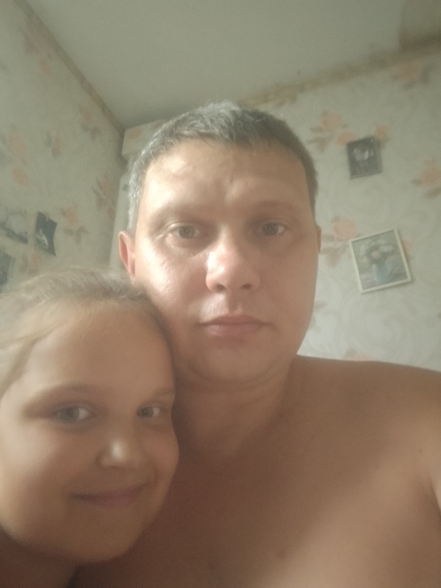 The media file is a selfie taken indoors, likely in a bedroom, with a man and a young girl positioned in the foreground. The man's face is partially obscured by the girl, who is leaning her head against his shoulder. The background features wallpaper with a floral pattern and two small framed pictures hanging on the wall. The lighting appears to be artificial, suggesting the photo was taken indoors, and there are no clear indicators of the time of day or weather conditions. The overall mood seems casual and intimate, possibly indicating a moment of familial affection. The image is slightly blurry, common in selfies. There is no visible text in the image. The location context provided is Pavlysh, Ukraine.