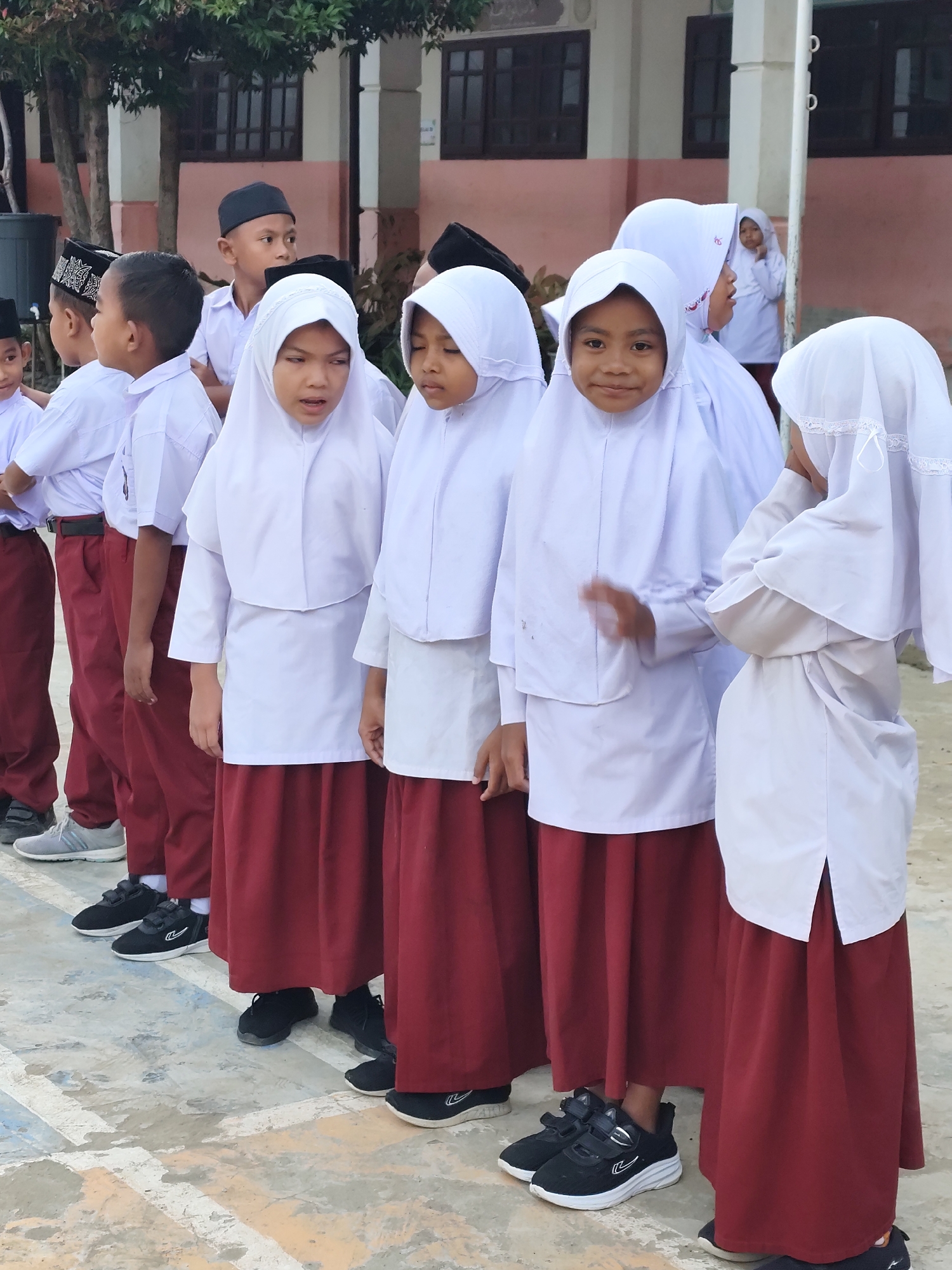 A group of young students, likely in uniform, stand in a line outdoors. Most are wearing white shirts and dark red skirts or pants, and headscarves. The students appear to be of elementary school age. In the background, there is a building with windows and some trees. The ground is paved with what looks like concrete. The overall scene suggests a school gathering or assembly.