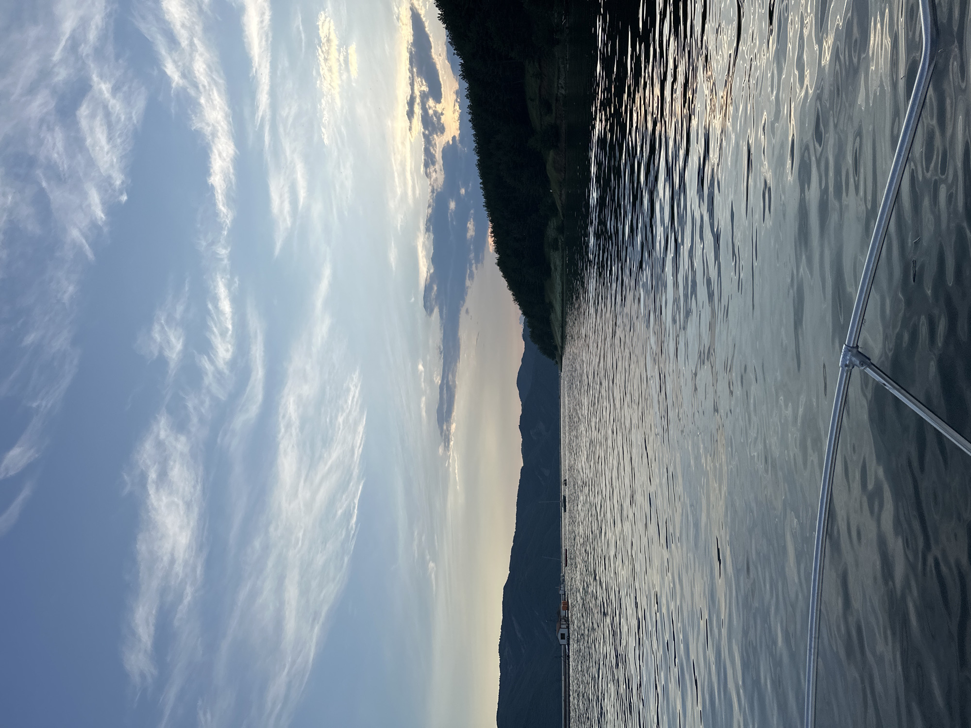 The image displays a wide shot of a tranquil lake or reservoir at dusk or dawn, likely situated near Brosh, Bulgaria, viewed from the bow of a boat. The upper third of the frame shows a pale blue sky with wispy, elongated cirrus clouds stretching horizontally. Towards the right horizon, the sky transitions to a muted gray-blue, with darker, more defined clouds obscuring a small patch of warm, orange-yellow light, indicative of the setting or rising sun.
The middle ground features a horizon line delineated by a range of dark, undulating mountains or hills covered in dense vegetation on the left. On the right, a closer, steeply sloped forested shoreline is visible, densely packed with dark green coniferous trees extending down to the water's edge.
On the water's surface in the mid-left, a small white building with a red roof, possibly a boathouse or hut, is situated on a pier or floating dock near the base of the distant mountains. Several small boats are moored or anchored nearby, including one with a visible mast suggesting it is a sailboat, and another smaller, reddish vessel. No individuals are discernible in the scene.
The lower two-thirds of the image depict the dark, rippled surface of the water, which reflects the subdued light from the sky and the surrounding landscape, showing subtle variations in tone. The very bottom foreground reveals a metallic, tubular rail, indicative of the bow rail of the vessel from which the photograph was captured.