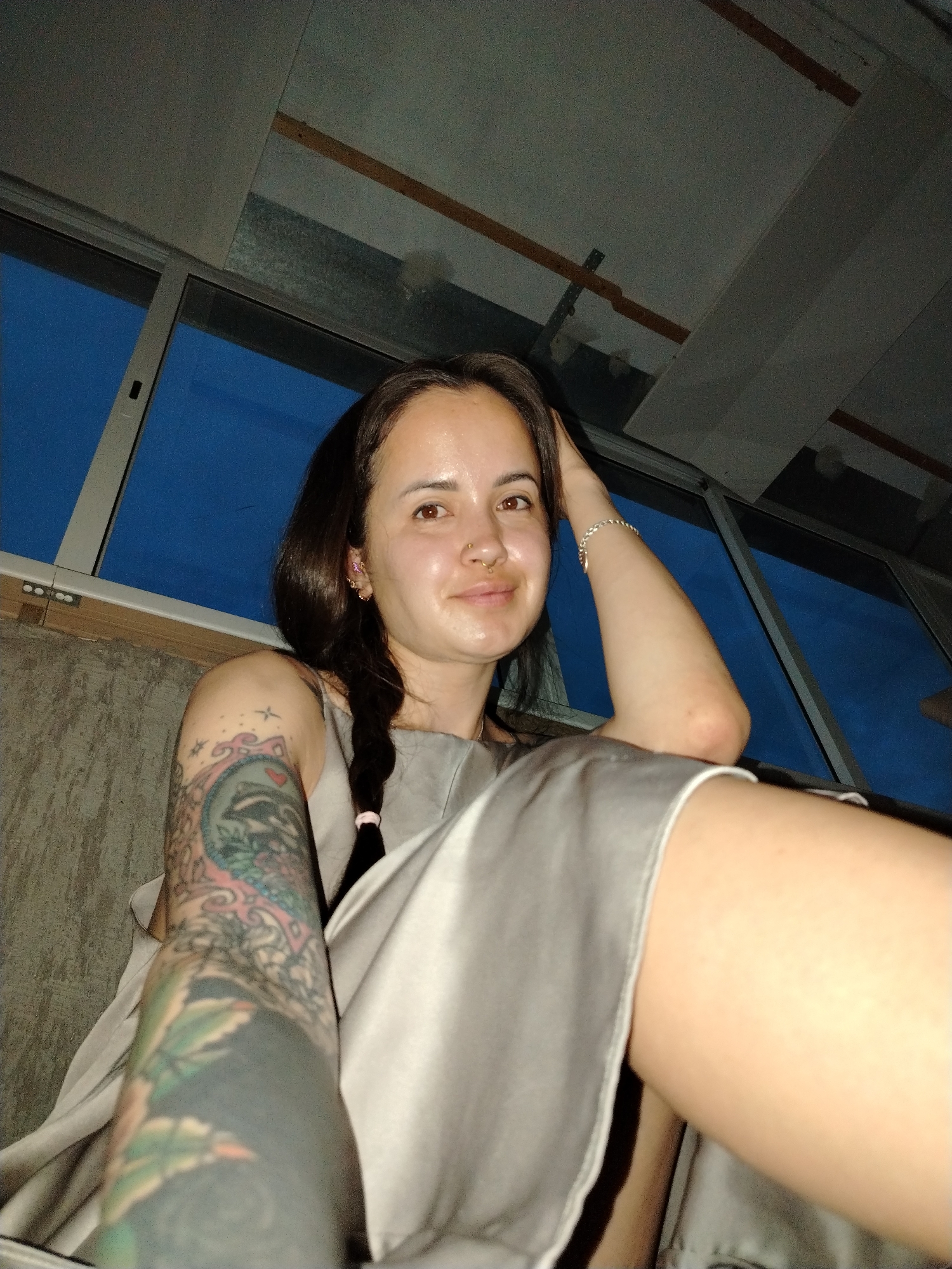 A woman is in an indoor setting, possibly in Noviy, Russia (based on city reference). She has dark hair in a braid with a pink hair tie, fair skin, and facial piercings (septum ring and earrings). She is wearing a light-colored dress. The woman has a tattoo sleeve on her left arm. The background shows a window with a dark blue outside view and a ceiling with wooden beams. She appears to be sitting or reclining.