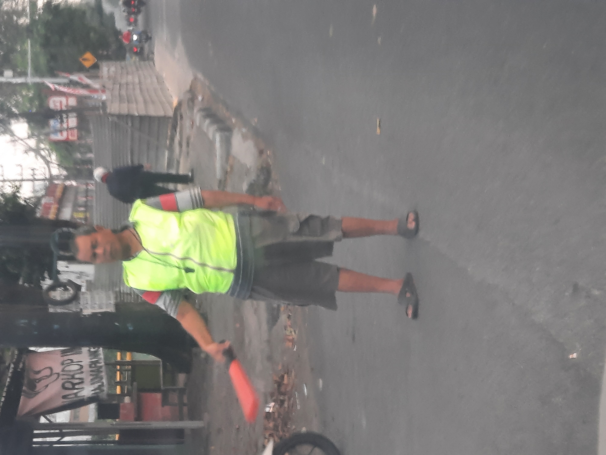 A man stands in the middle of a street in Banjarkemantren, Indonesia, holding a red signaling paddle. He is wearing a neon yellow vest over a grey shirt with red and grey stripes on the sleeves. He has on grey cargo shorts and dark flip-flops. The man has dark hair styled in a mohawk.
Behind him, a banner with Indonesian text, including "NARKOP IN" and "MAJU BANGUN," is hanging from a structure. To the right of the man, a staircase leads up to a raised area. Several motorcycles are visible on the street in the background, with one rider in a red shirt. The street is paved, with a curb and sidewalk on the right side of the image. The scene appears to be outdoors, possibly in an urban or semi-urban area. The lighting suggests it may be dusk or early morning.