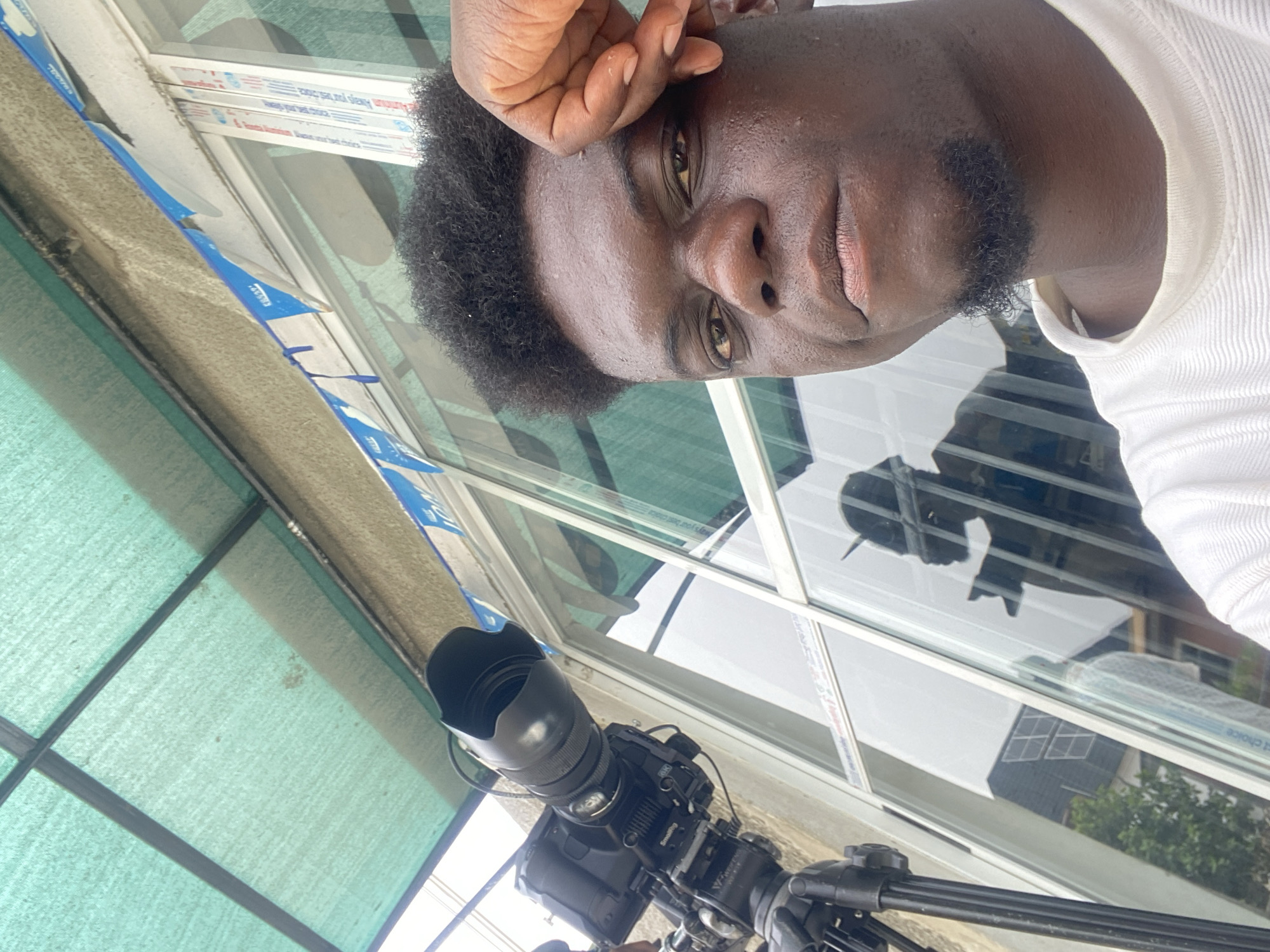 A close-up shot of a man with dark skin and a black beard, wearing a white t-shirt. His right hand is touching his forehead. The background includes a window with reflections. A camera on a tripod is visible below. The location is in the city of Unknown, Unknown.