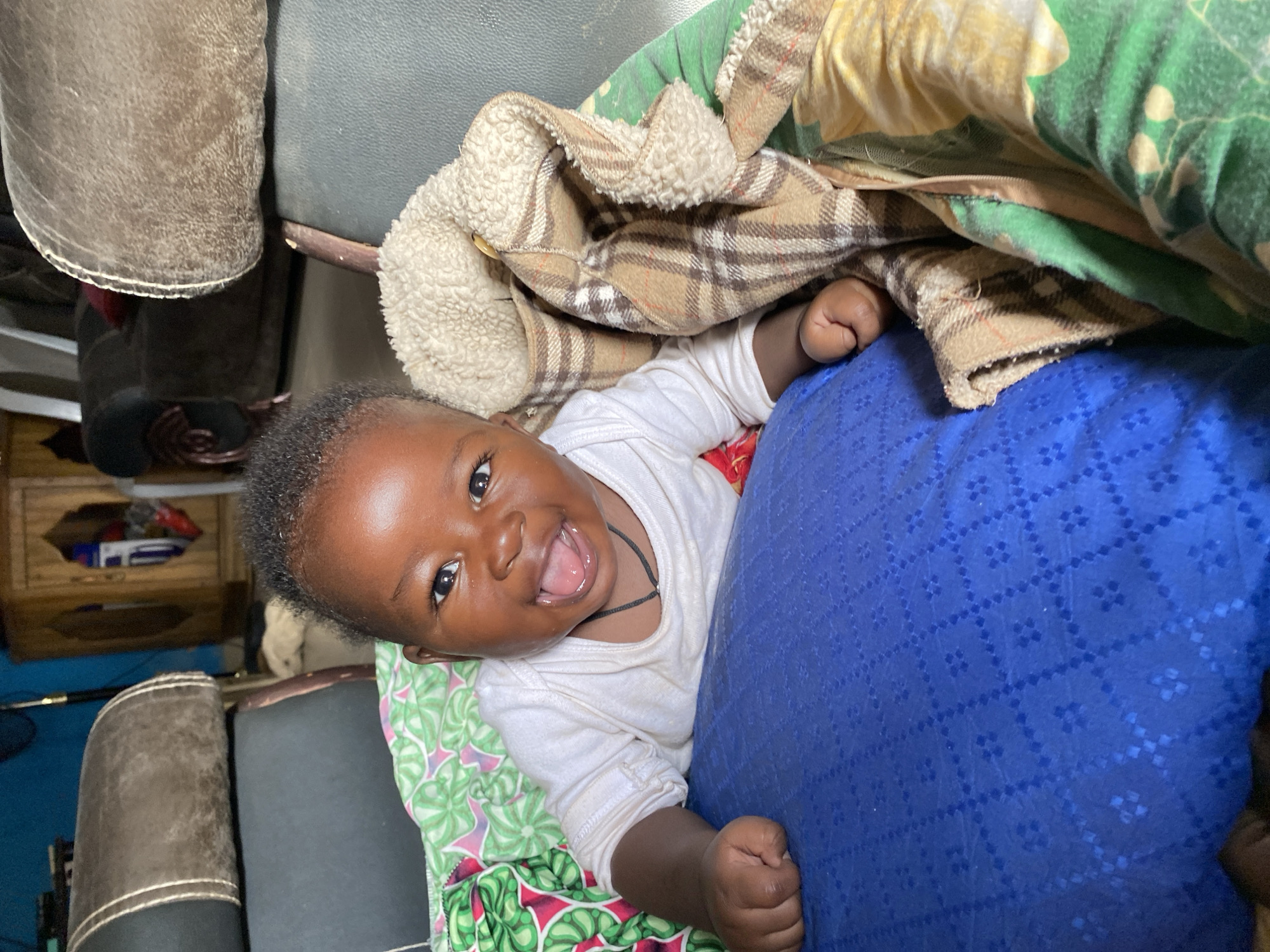 A joyful baby, appearing to be an infant or young toddler, is captured in a close-up, angled shot, looking directly at the camera with a wide, open-mouthed smile. The baby has dark skin, bright eyes, and short, dark curly hair, conveying a sense of happiness and playfulness. A thin dark cord or necklace is visible around the baby's neck. The baby wears a plain white long-sleeved top.
The baby is positioned amidst a collection of soft furnishings. On the left side, a vibrant green and pink patterned fabric, possibly a blanket or sheet, is visible. To the baby's right, a textured dark blue fabric, likely a pillow or cushion, supports the baby. Draped over the upper right side of the frame is a brown and white plaid blanket with a cream-colored, fuzzy lining, partially covering the baby. Beneath this, a green and yellow patterned fabric is also visible.
In the background, elements of upholstered furniture in shades of brown and dark grey suggest an indoor setting, likely within a home. The overall scene radiates warmth and contentment, capturing an endearing moment of the baby smiling. The image provides no visible text. The location context is Vom, Nigeria.