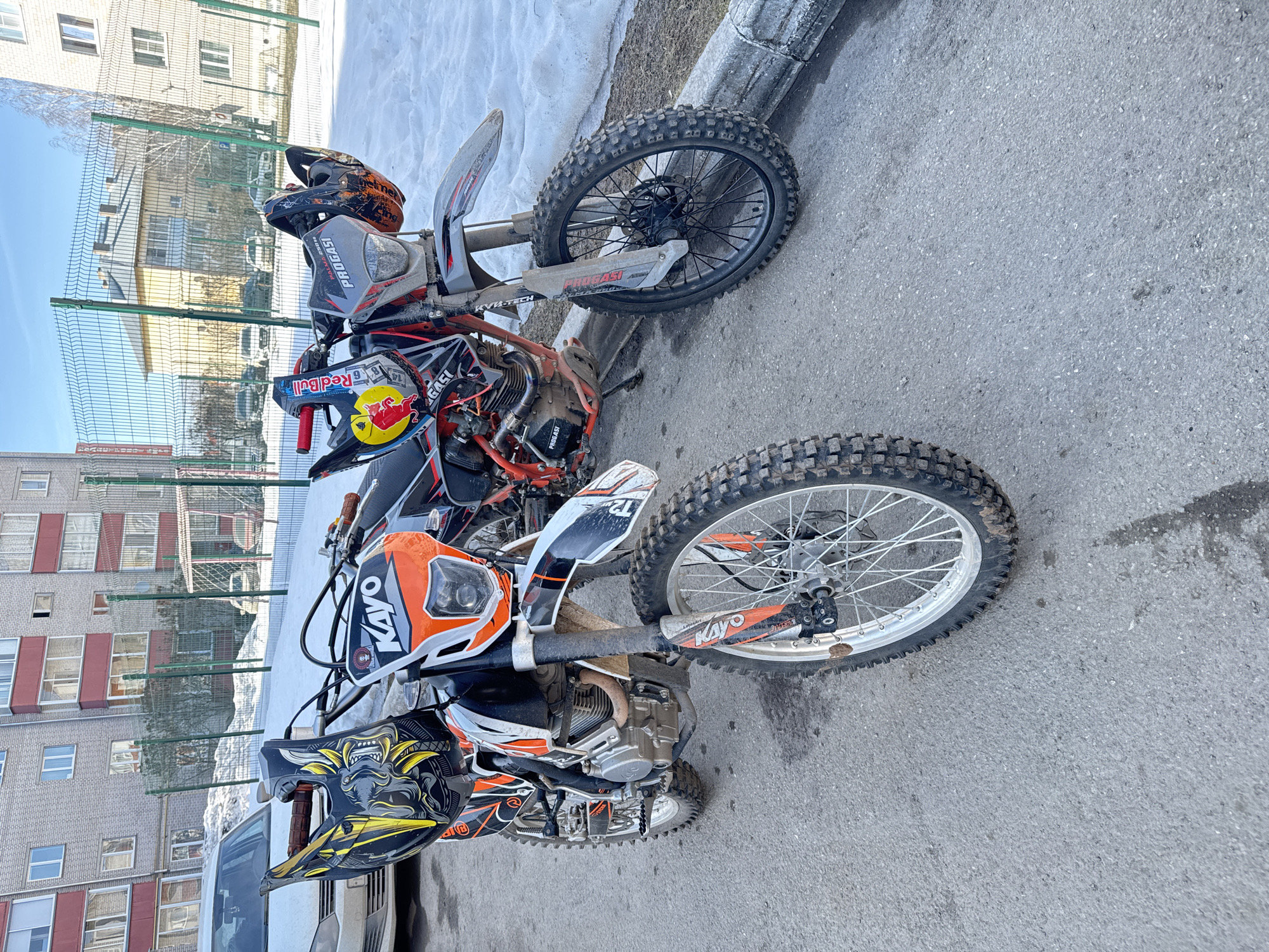 The image shows two dirt bikes parked on an asphalt surface near a curb. In the background is a multi-story brick building with a green mesh fence in front of it. There is also some snow visible near the curb. The front dirt bike is an orange and white Kayo motorcycle with a black and yellow helmet resting on the handlebars. The rear dirt bike is red, white, and black with a Red Bull logo and an orange helmet on the seat. The location is inferred to be in the city of Zav'yalovo, Russia.
