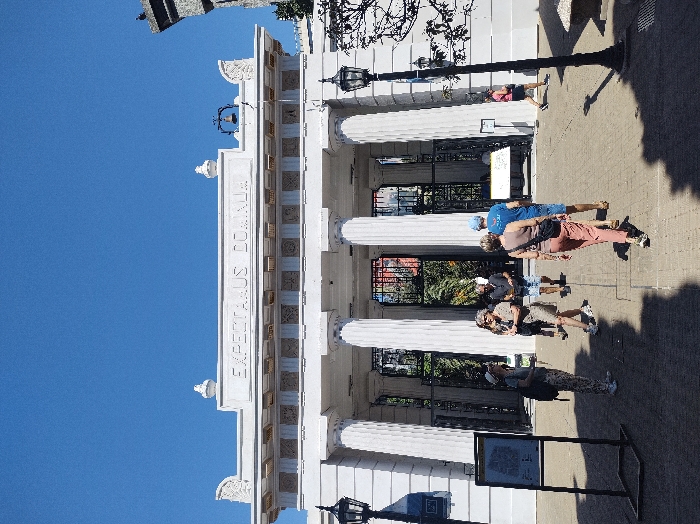 A low-angle shot presents a white building with prominent columns under a clear blue sky. The building's facade features the inscription "EXPECTAAUS DU" along the top, complemented by decorative details. Several people are positioned in front of the building, some standing and others walking. A lamppost stands near the building, casting a shadow on the paved ground. The composition suggests a tourist or historical site.
