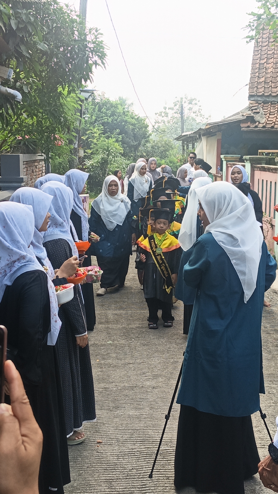 The image captures a graduation procession taking place on a concrete pathway in Tigaraksa, Indonesia. A young boy, adorned in a graduation cap and gown with a sash that reads "PAUD Islam Tigaraksa," is the central figure, walking forward. He is surrounded by a group of women, most of whom are wearing hijabs. Some of the women are holding bowls filled with flower petals, suggesting a celebratory event. The background shows residential buildings with tiled roofs and lush green trees, indicating a typical neighborhood setting. The overall atmosphere appears festive and communal, with people gathered to witness the graduation.