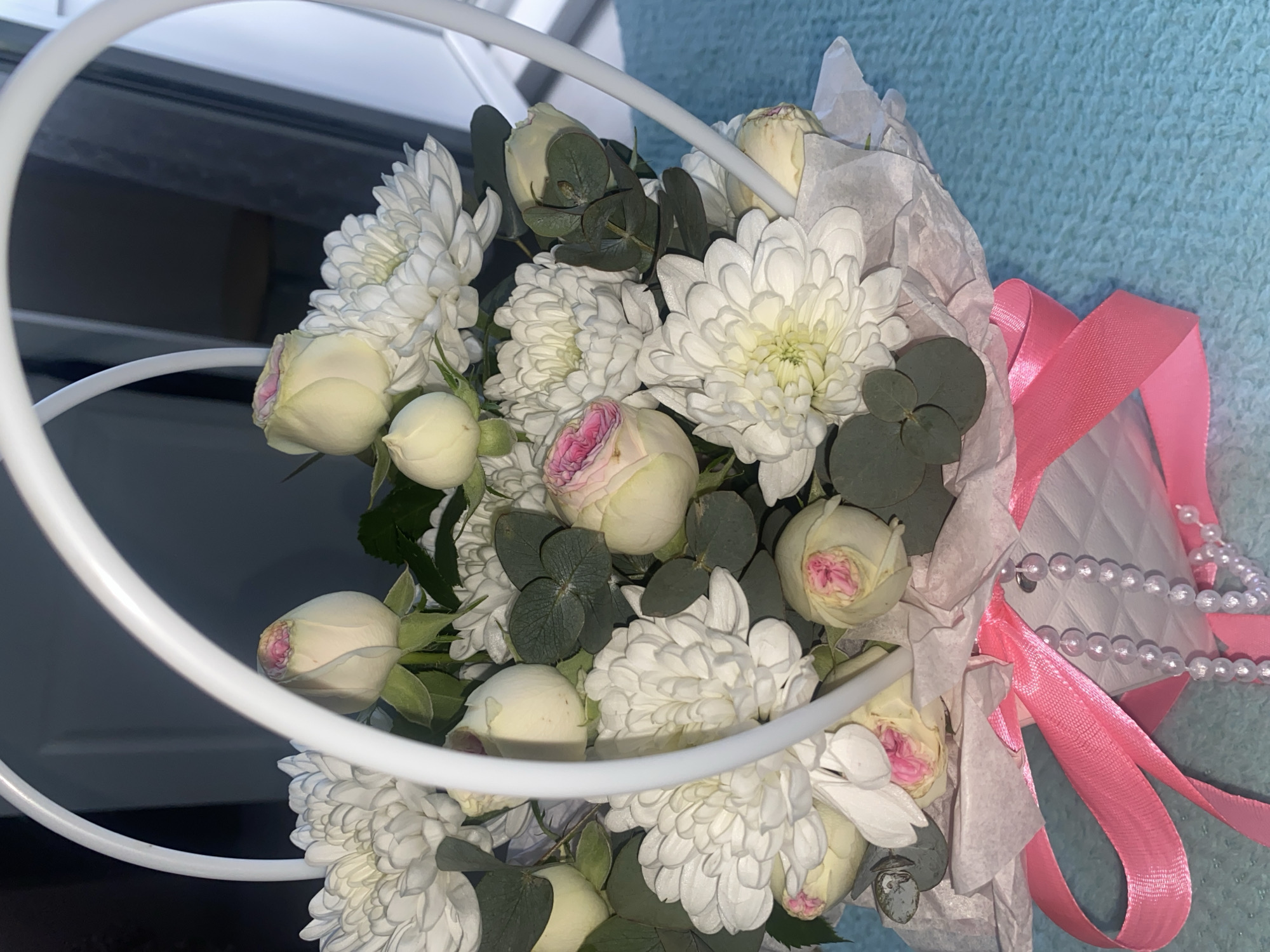 A close-up shot displays a bouquet of white chrysanthemums and roses, wrapped in white paper and tied with a pink ribbon. The bouquet also features eucalyptus leaves and a string of white beads. The arrangement is partially contained within a white, circular metal frame, potentially a decorative holder. The background is a mix of light blue and dark gray, suggesting an interior setting with varying light conditions. The image doesn't provide sufficient information to confirm its location as Izhevsk, Russia.