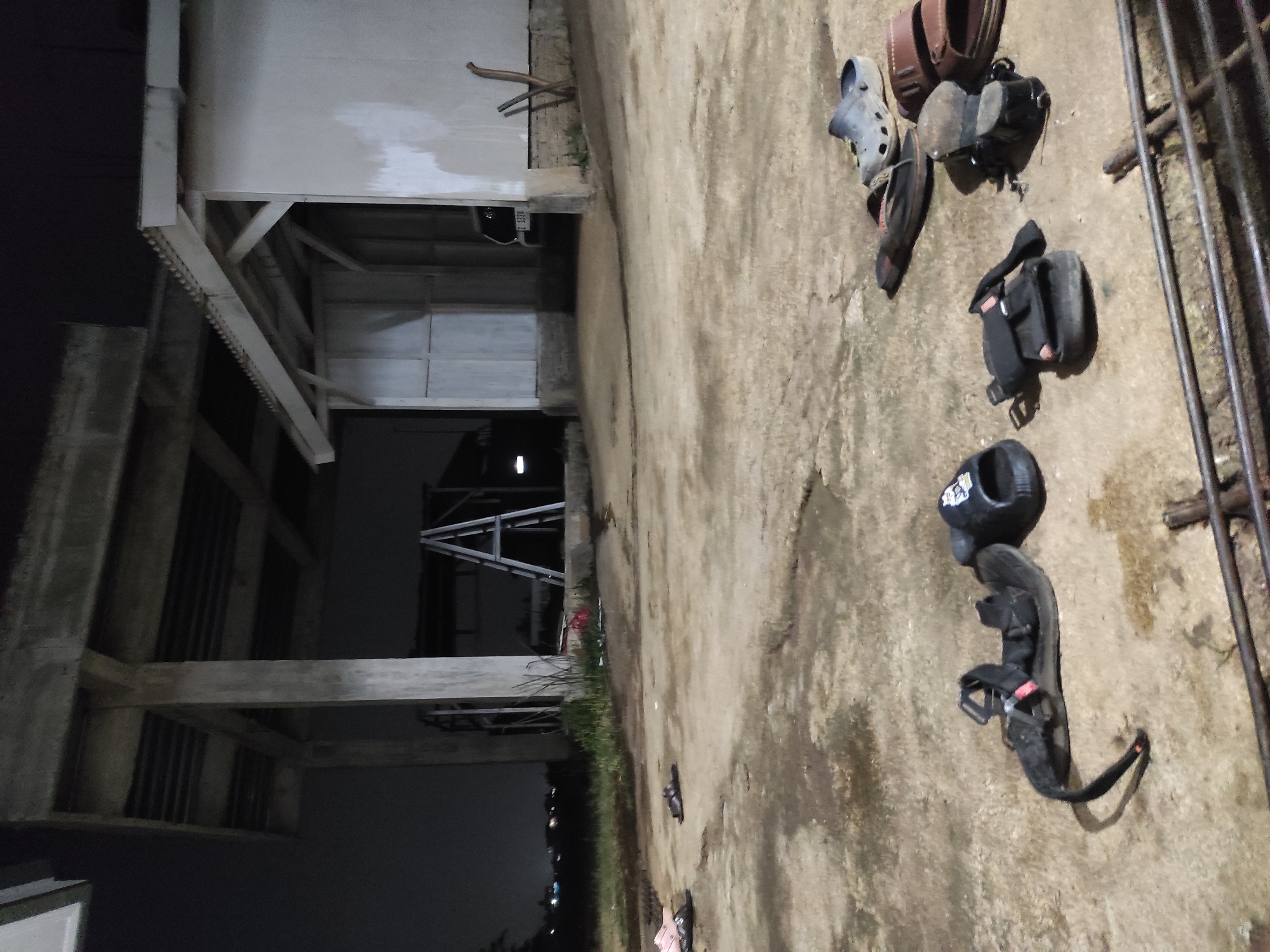 The image depicts an outdoor scene at night, likely in Sumedang, Indonesia, characterized by a concrete ground with several pairs of scattered footwear. In the foreground, a collection of sandals and flip-flops are visible. These include black sandals with straps, a pair of brown sandals, and some dark-colored flip-flops. In the background, there are structures that appear to be a covered outdoor area supported by concrete pillars, and a building with a white wall and a corrugated roof. A ladder is propped up against one of the structures. The environment appears somewhat unkempt, with patches of grass and debris visible. The lighting suggests it is nighttime, with artificial light illuminating the area. The overall impression is of an informal outdoor space, possibly a residential or communal area, where shoes have been left casually.