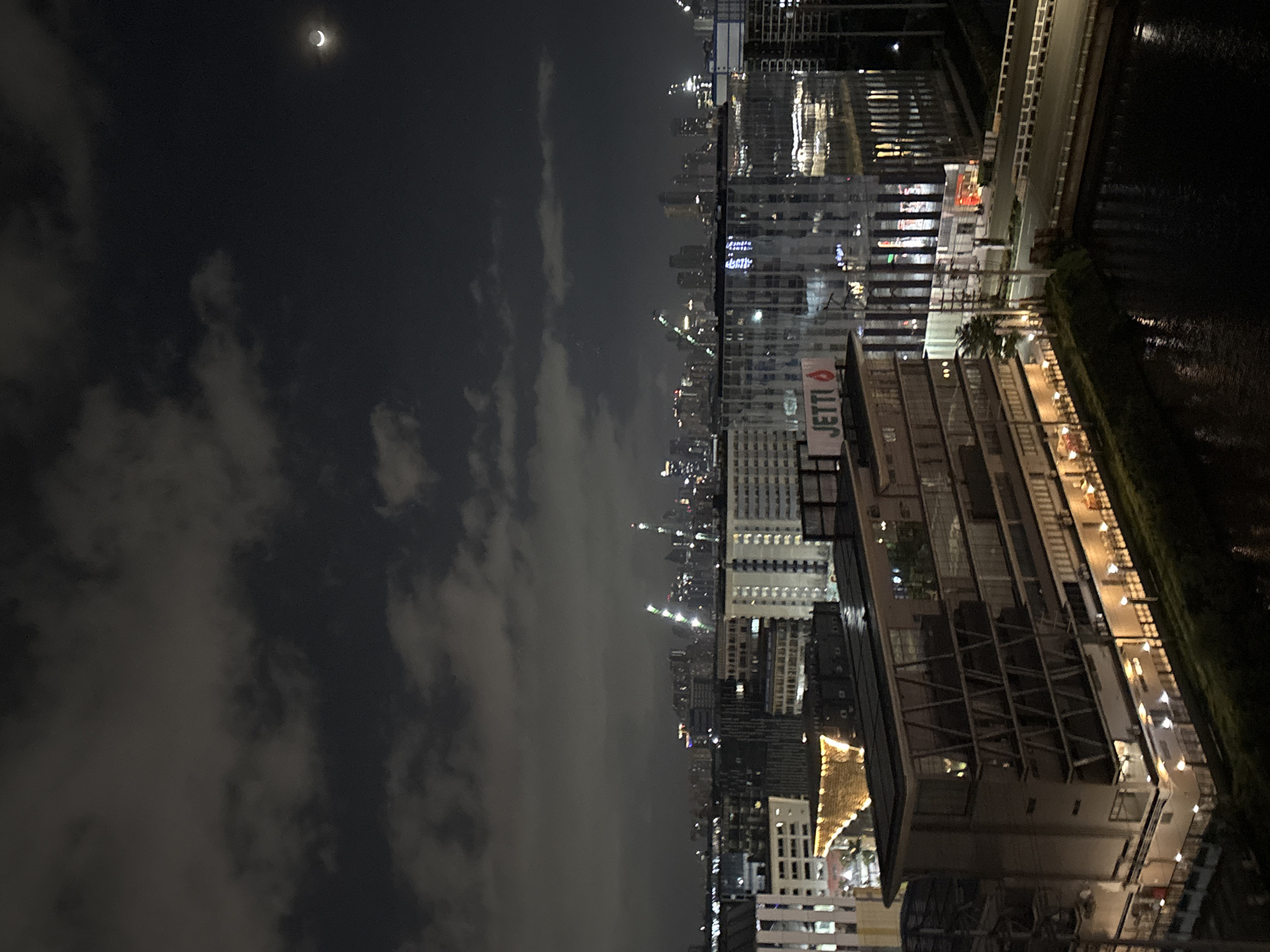 The image depicts an urban night scene in Pasay City, Philippines. In the foreground, a dark body of water, likely a river or canal, is visible. A bridge spans the water, connecting to a road. To the left of the water and bridge is a multi-story building with glass windows, featuring prominent lighting at its base and illuminated floors. A sign with the word "JETTI" is affixed to the side of this building. Behind this structure, a cityscape stretches into the distance, characterized by numerous tall buildings and skyscrapers. Construction cranes, illuminated with green lights, are visible among the buildings, suggesting ongoing development. The sky is dark, with scattered clouds, and a sliver of a crescent moon is present in the upper right quadrant of the image. The overall impression is that of a bustling, modern urban environment at night.