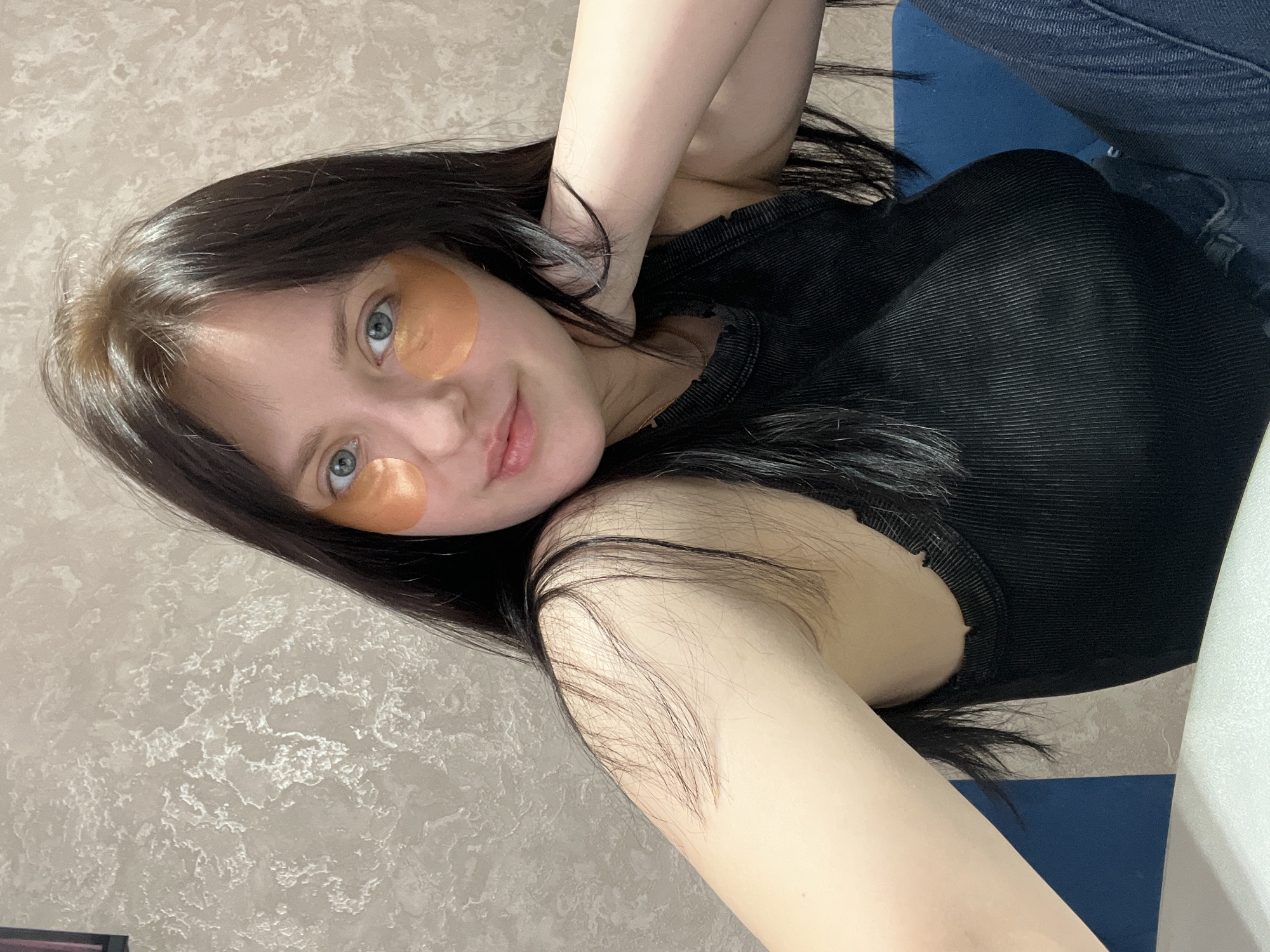 A young woman with long dark hair poses for a selfie indoors. She has light skin, blue eyes, and is wearing what appear to be orange under-eye masks. She is wearing a dark-colored, ribbed tank top and denim jeans. Her left arm is raised and positioned behind her head. The background consists of a textured light beige wall, a blue surface, and a partial view of a white object. There is no information to connect the location to Марковский, Russia.
