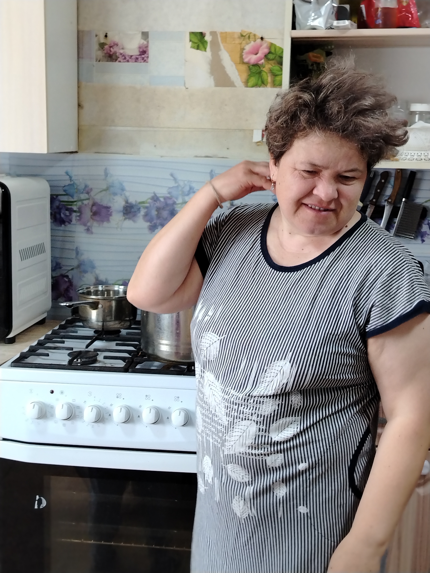 A woman stands in a kitchen next to a white stove with two pots on it. The woman has short, curly brown hair and is wearing a striped dress. She has her hand raised to her neck, and she is smiling. The kitchen has cabinets and drawers, and a wall with floral wallpaper.