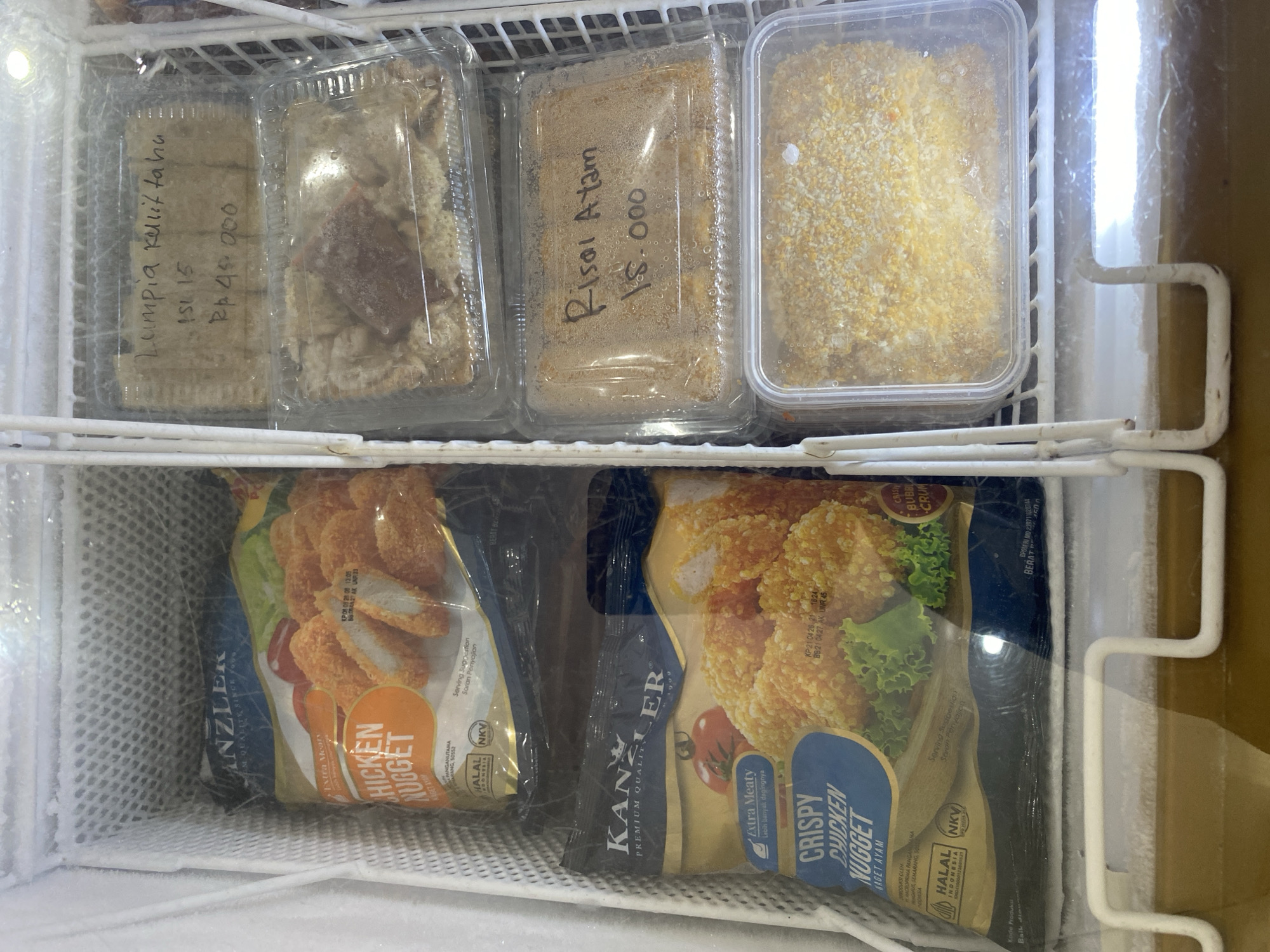 This is a close-up, eye-level photograph of the interior of a commercial freezer, likely in a store or restaurant in Payatieng, Indonesia. The freezer is filled with various frozen food items, predominantly chicken nuggets and what appear to be spring rolls or similar pastries.
On the left side of the freezer, there are two large, individually packaged bags of "KanZLER Premium Quality Chicken Nugget". The bags are primarily blue and yellow with images of chicken nuggets. One bag has text indicating "Extra Meaty" and "Crispy Chicken Nugget". Both bags display Indonesian language text, including "Halal Indonesia".
On the right side of the freezer, there are several clear plastic containers holding food items. The top container has handwritten text that reads "Lumpia Kulit Tahu Asli 15 Rp 45.000". The second container down has handwritten text that reads "Risoi Ayam 15.000". These containers hold what appear to be filled pastries, with some showing signs of being frozen solid. The third container from the top, partially visible at the bottom right, also holds a breaded frozen food item.
The freezer's interior is frosted, indicating its operational state. The shelves are white plastic, and the overall environment is cold and industrial. There are no people or animals visible in the image. The lighting appears to be from within the freezer itself, casting a cool, artificial glow on the products.