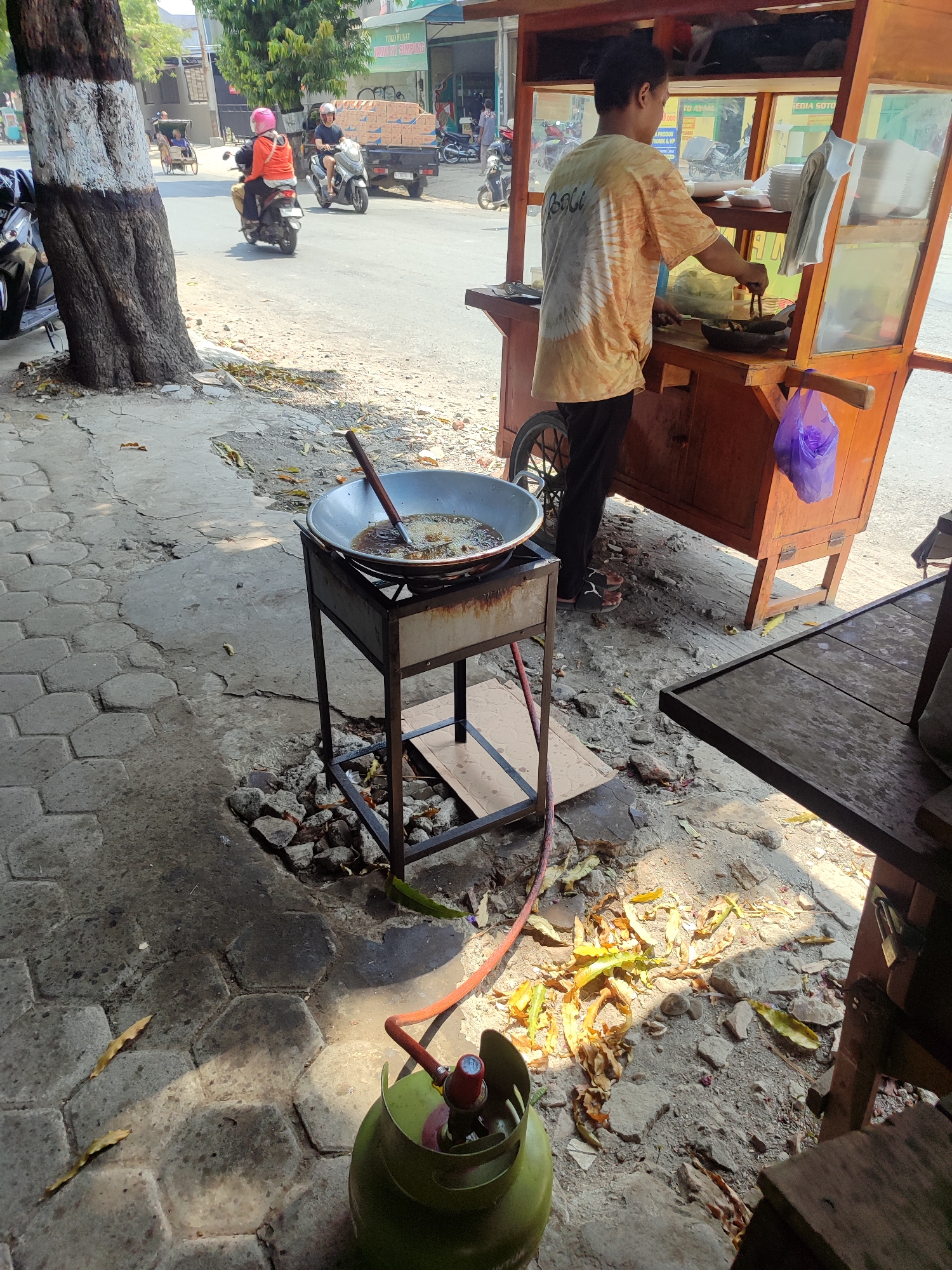 The image depicts an outdoor scene at a street food stall in Kagok, Indonesia. A person, wearing a yellow and white tie-dye t-shirt and dark pants, is actively cooking at a portable food cart. The cart, made of wood with a glass display case, is positioned on the right side of the frame. A large wok filled with cooking oil sits on a metal stand in front of the cart, with a wooden cooking utensil resting inside. A green gas cylinder with a red regulator and hose is visible on the ground in the foreground, connected to the cooking apparatus. The ground is paved with hexagonal stones and littered with fallen leaves and debris. In the background, a street is visible with several vehicles, including motorcycles and a truck, suggesting a moderately busy urban environment. A large tree with a white-banded trunk stands on the left side of the image, casting shadows. The overall impression is of a typical bustling street food vendor operating in a localized Indonesian setting.