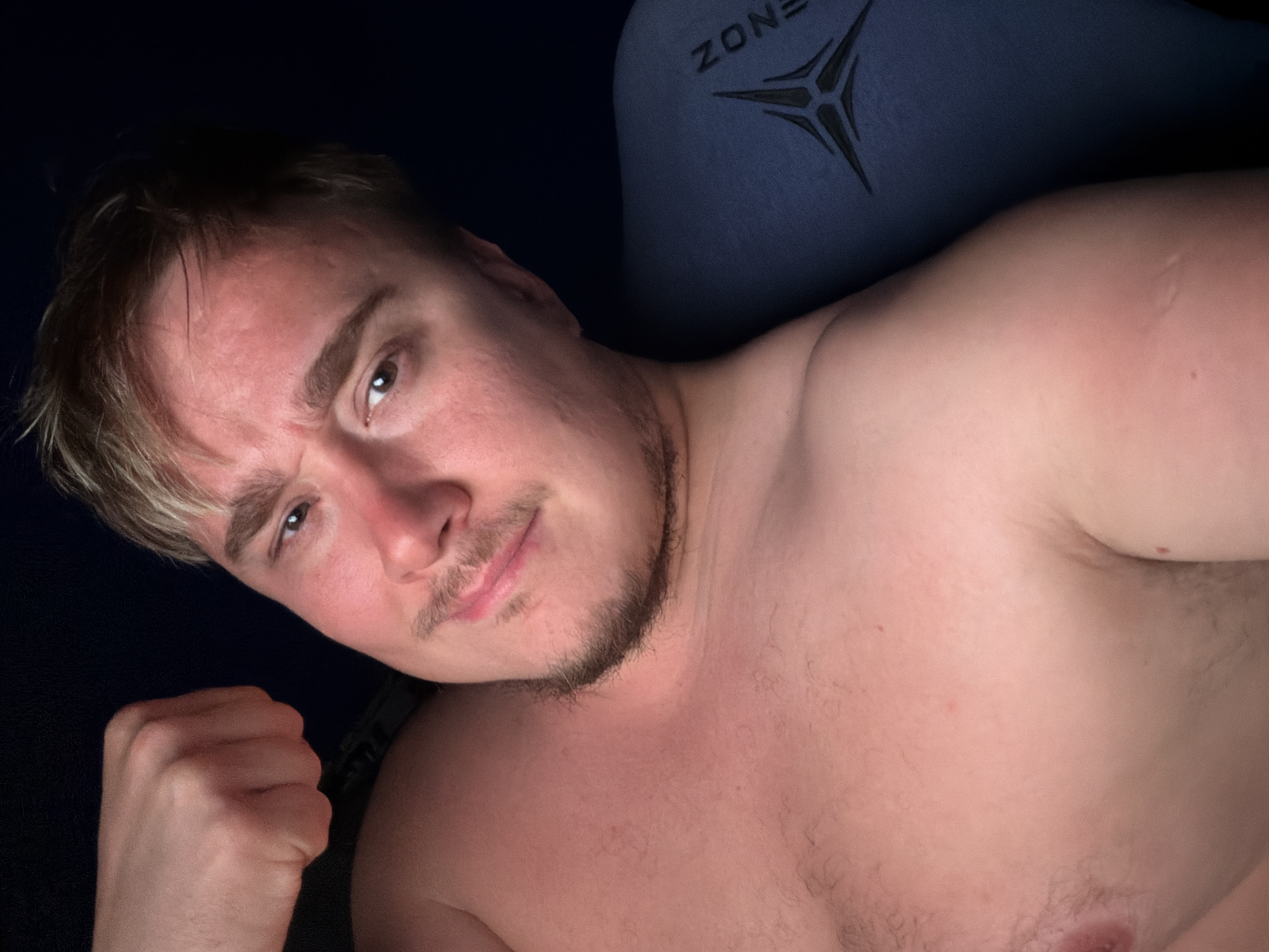 A young man, likely in his late teens or early twenties, poses for a selfie indoors, leaning back against a blue gaming chair. He has light skin, light brown hair with blonde highlights at the front, a mustache, and a short beard. He is shirtless, revealing his chest and shoulders, which have some light body hair. His left hand is clenched into a fist and held up near his shoulder.
The man looks directly at the camera with a determined or slightly mischievous expression, characterized by a raised left eyebrow and a subtle smirk. The setting appears dimly lit, suggesting nighttime or a darkened room, with a primary light source illuminating his face and upper body.
Behind him, the headrest of a blue gaming chair is visible, bearing a black logo and the partially discernible word "ZONE". The logo is a stylized three-pronged star or claw mark design. The image captures an intimate moment in what appears to be a personal space, such as a bedroom or gaming room, located in Chaikovsky, Russia.