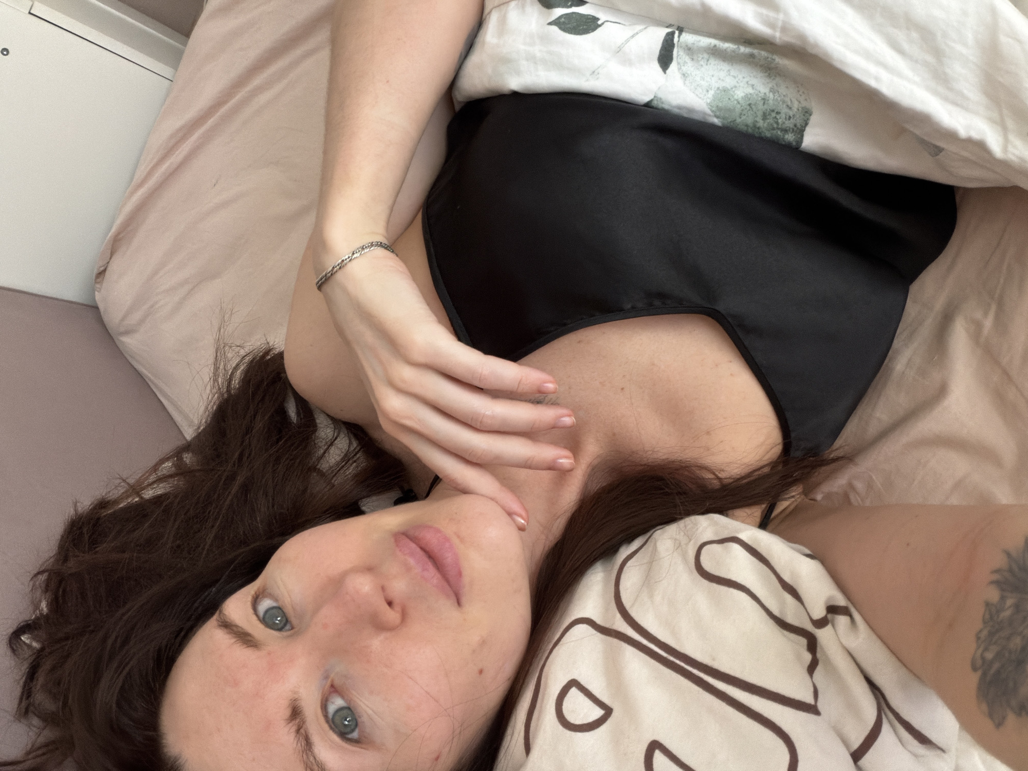 A woman lies in bed, taking a selfie from an overhead perspective, looking directly into the camera. She is positioned diagonally across the frame, with her head towards the lower left and her body extending towards the upper right. Her dark, wavy hair frames her face. She has light blue eyes and a neutral, relaxed expression, with her lips slightly parted.
She is wearing a simple black satin-like top or camisole. Her left arm is visible, bent at the elbow, with her hand near her face. A silver braided bracelet is worn on her left wrist. A faint tattoo is partially visible on her right arm, just below the elbow, peeking out from under the duvet.
The bedding consists of a pale pink or beige fitted sheet, a white duvet with a subtle green-grey floral or abstract pattern, and another pillow or duvet cover with a brown abstract, wavy line pattern. The background suggests a simple indoor setting, likely a bedroom in Perm, Russia, with a light-colored wall or headboard visible in the upper left corner. The lighting suggests it is daytime, with soft ambient light. There is no visible text in the image.