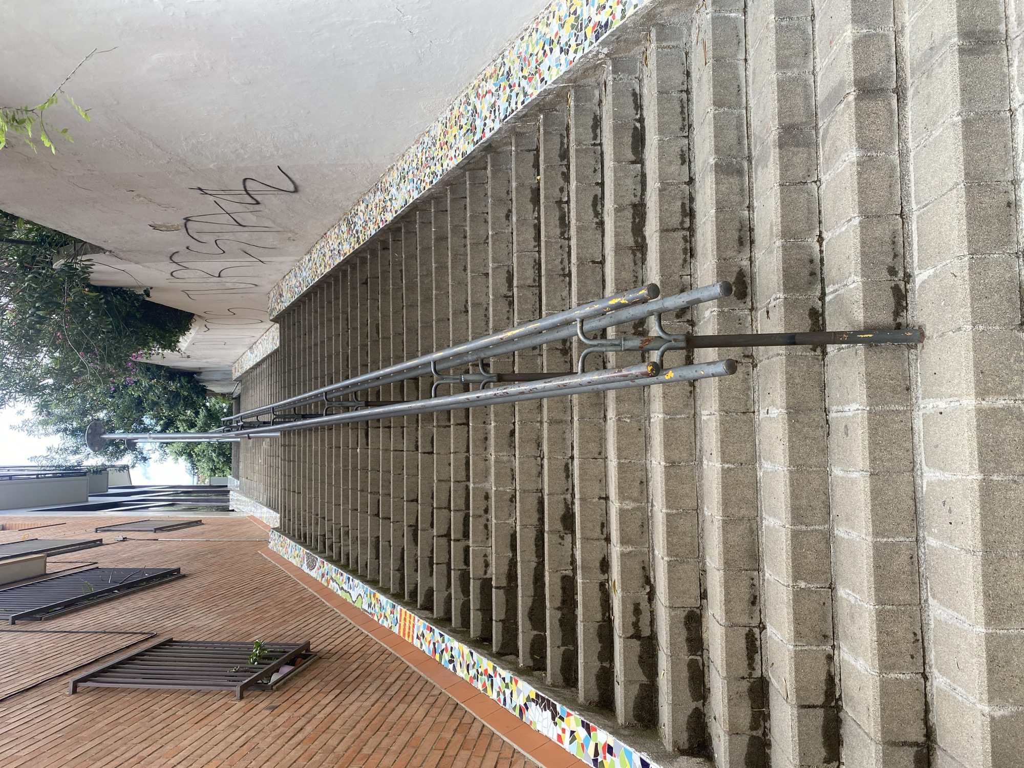 The image displays an outdoor architectural setting, characteristic of an urban environment, likely in Barcelona, Spain. The central feature is a wide concrete staircase or ramp, ascending diagonally from the lower left to the upper right. The steps are constructed from large, textured concrete blocks, and alongside them runs a metal handrail, which appears aged with visible wear and patches of peeling yellow paint.
A distinctive decorative element is a vibrant border of mosaic tiles that lines the base of the concrete structure and an adjacent lower wall. These irregularly shaped tiles feature a rich palette of colors including red, yellow, blue, green, white, and black. Below this mosaic, a red-tiled ground area extends, on which several dark metal grate-like structures are visible, possibly serving as covers for planters or ventilation.
An off-white, concrete or stucco overhead structure extends above the staircase. The underside of this surface is marked with black, stylized graffiti, appearing as an illegible tag with several abstract forms. To the far left of the scene, beyond the architectural elements, lush green trees and bushes are visible, adding a natural touch to the landscape.
The photograph captures a static, daytime scene, likely under indirect light or overcast conditions, as indicated by the soft shadows. No people or specific activities are depicted. The composition highlights a blend of utilitarian construction materials, decorative artistic elements, and urban street art.