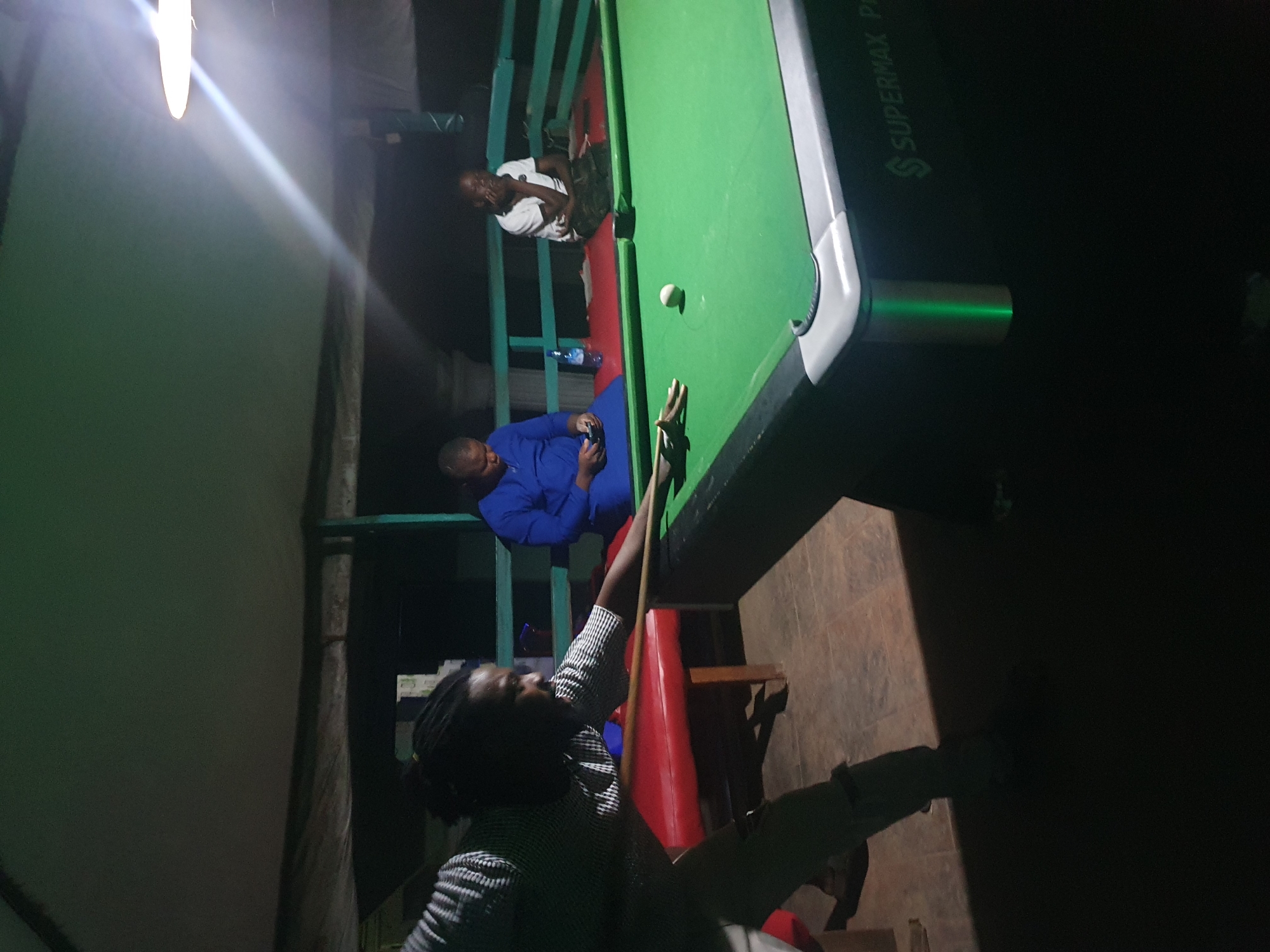 The image depicts an indoor billiards scene at night, possibly in Lokoja, Nigeria. Three individuals are present. In the foreground, a man with braided hair, wearing a patterned shirt and light-colored pants, is actively playing billiards, poised to strike a ball with a cue stick. He is positioned on the left side of the frame. To the right of him is a green billiards table with a white cue ball visible.
In the mid-ground, a person dressed in a blue garment is seated on a red cushioned bench behind the billiards table, looking down at a mobile phone. Further to the right, another individual is seated on the same bench, wearing a white t-shirt and dark pants, with their hand resting near their face.
The environment appears to be an open-air covered area, suggested by the visible ceiling structure and the surrounding dimly lit background, which includes wooden railings and some vegetation. A bright light fixture hangs from the ceiling, illuminating the scene. The floor is tiled. A bottle of water is visible on the bench between the two seated individuals. The billiards table has "SUPERMAX PRO" branding on its side. The overall lighting is dim, with the primary light source being the overhead lamp.