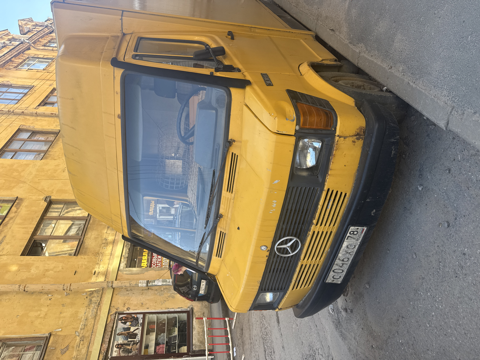 The image shows a yellow Mercedes-Benz van parked on a cobblestone street in Saint Petersburg, Russia. The van is a box truck with a high roof and a visible Mercedes-Benz logo on the front grille. The license plate is partially visible and reads "C046XC 78 RUS". In the background, there is a weathered yellow building with multiple windows and a shopfront. A black car with a visible license plate is parked behind the van. The street appears to be a city street with a sidewalk and some debris on the ground. The scene is likely a residential or commercial area given the building and shopfront.