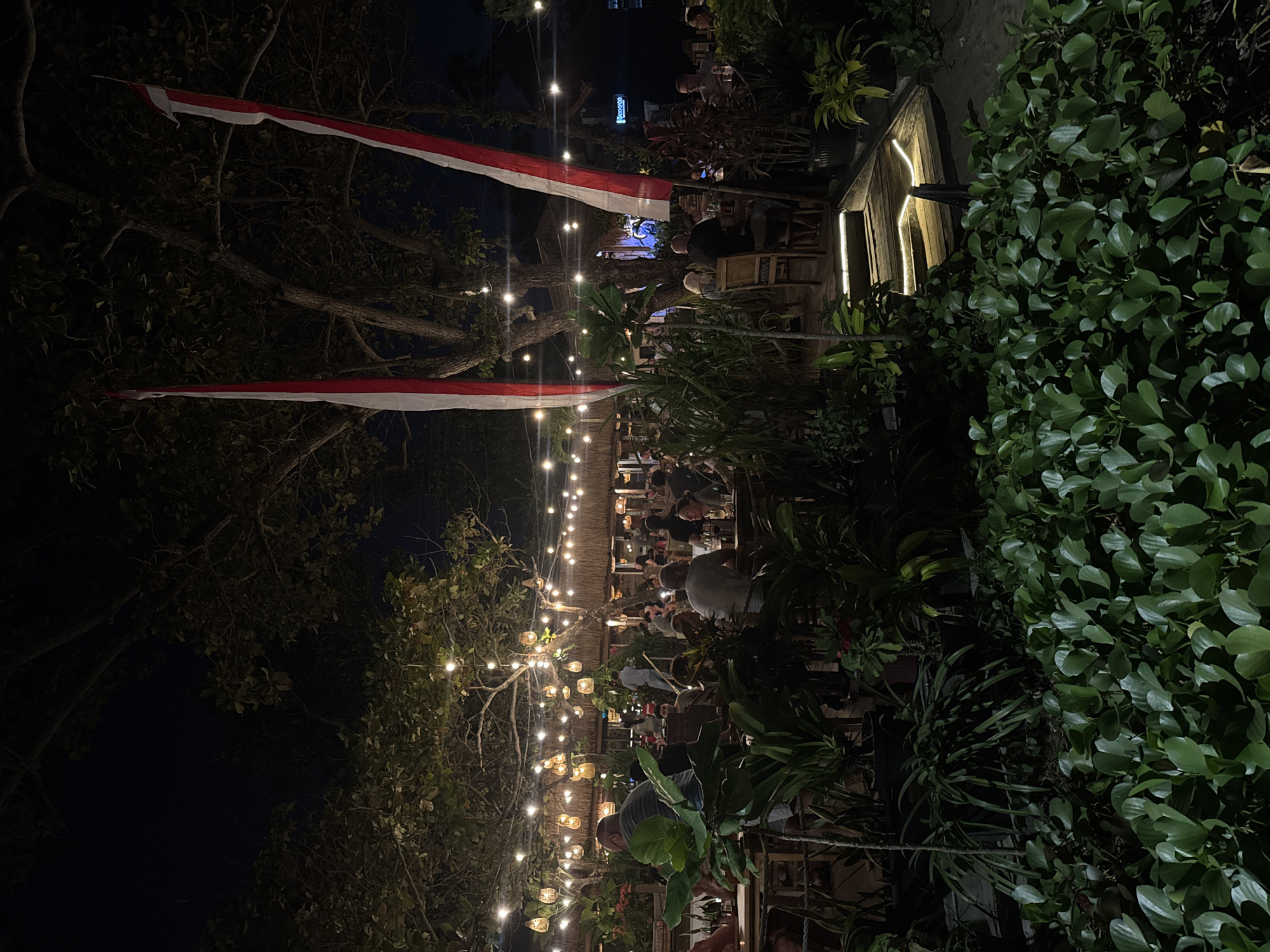 A nighttime outdoor dining scene is depicted in Kuta, Indonesia, with numerous patrons seated at tables under a canopy of trees adorned with strings of warm-toned lights and woven pendant lamps. Two tall red and white flags, likely the Indonesian flag, are prominently displayed. The foreground is filled with lush, green foliage. In the mid-ground, people are dining and conversing at wooden tables and chairs. Beyond the dining area, a thatched-roof building with more lighting and patrons is visible. A wooden staircase illuminated by soft lights leads up towards the dining area from the lower right of the frame. The overall atmosphere suggests a relaxed and lively restaurant setting on a beachside location.