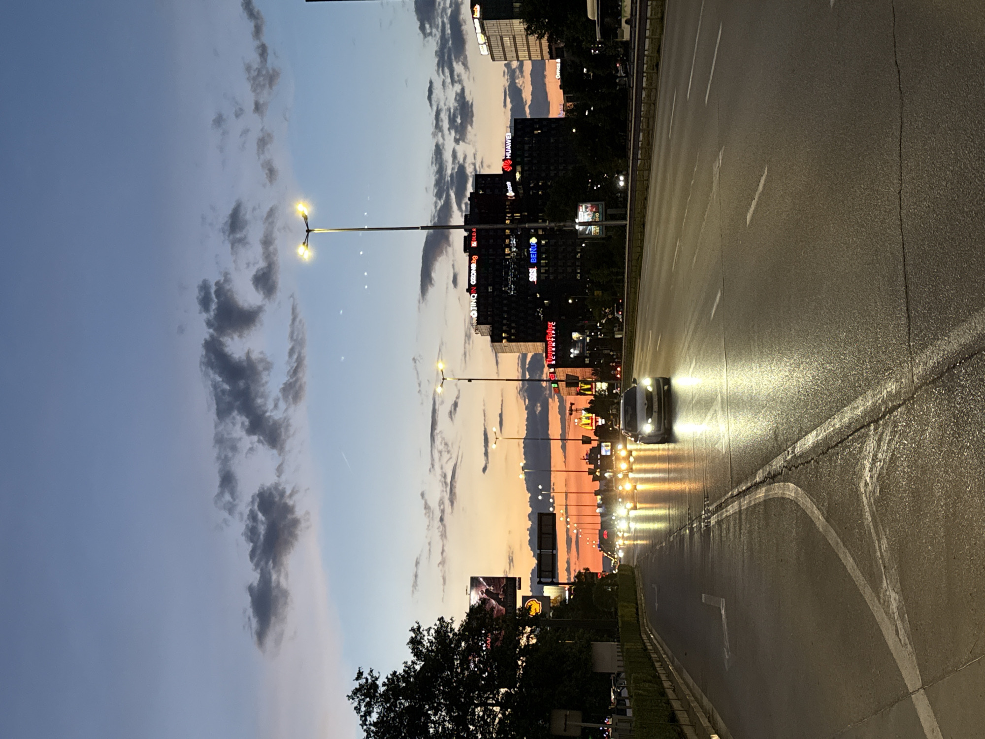 The image depicts a city street in Sofia, Bulgaria, at dusk. The sky is a gradient of soft blues and oranges, with scattered clouds. On the right side of the frame, a multi-lane road is visible, with cars driving and their headlights illuminated. The road surface appears wet, suggesting recent rain. Streetlights line the left side of the road, casting pools of yellow light.
In the background, several tall buildings are illuminated with various colored lights, some displaying text. Visible text includes "HUAWEI" and "MCDONALD'S". The overall atmosphere is one of a bustling urban environment transitioning from day to night, with a somewhat moody and dramatic sky. The composition is vertical, making it appear as if the scene is upside down. The image captures a typical evening scene in a city, with traffic, lighting, and architectural elements all contributing to the urban ambiance.