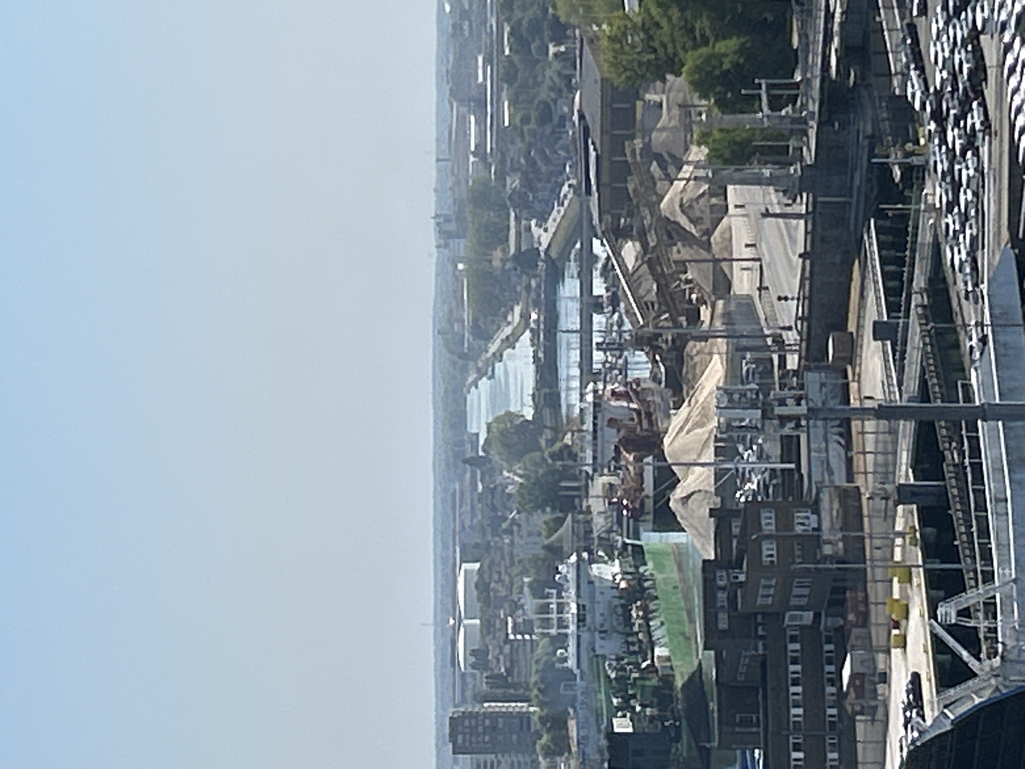 The image is an aerial view of the port of Ostend, Belgium, on a clear, sunny day. The foreground shows a large, multi-lane road with numerous parked cars. Further back, a large industrial area is visible, with various buildings, cranes, and piles of sand or gravel. A large green ship is docked in the harbor, with several other vessels visible in the background. A canal or waterway cuts through the city, with bridges spanning it. In the distance, more buildings and industrial structures can be seen, extending towards the horizon. The sky is a pale blue and cloudless. The overall scene depicts a busy, functional port environment.