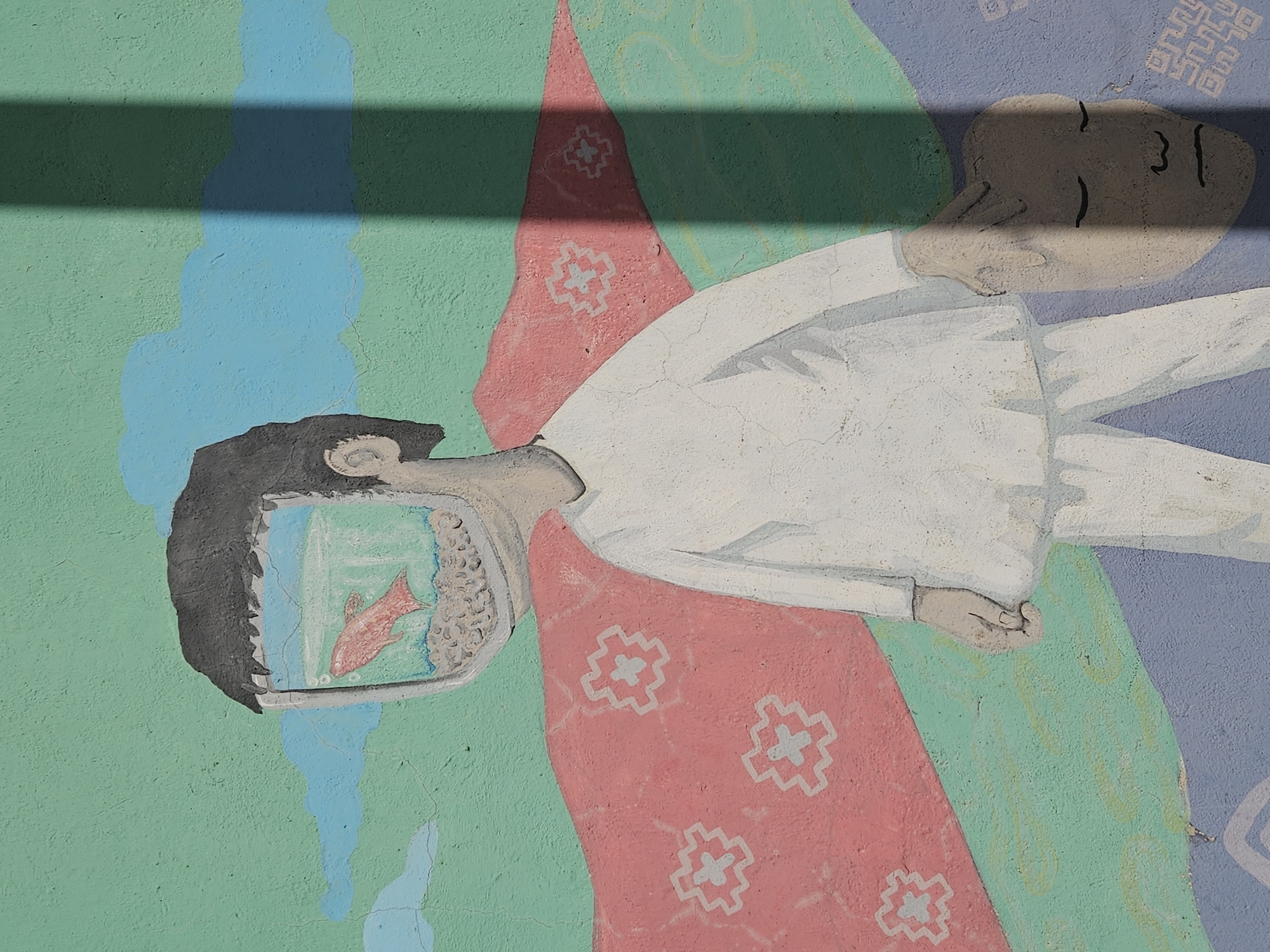 The image is of a mural, likely located in Osorno, Chile, showing a figure lying horizontally against a painted backdrop of blue, green, and lavender. The figure is painted in shades of white and grey, wearing what appears to be a long-sleeved garment. The head of the figure is depicted as an open television screen displaying a red fish swimming in a blue, watery environment. The rest of the figure appears to be a regular human head. The painted backdrop behind the figure includes geometric patterns and abstract color fields. The mural's surface has cracks and imperfections, typical of an outdoor wall. A dark shadow passes across the image, obscuring the upper section of the painting.