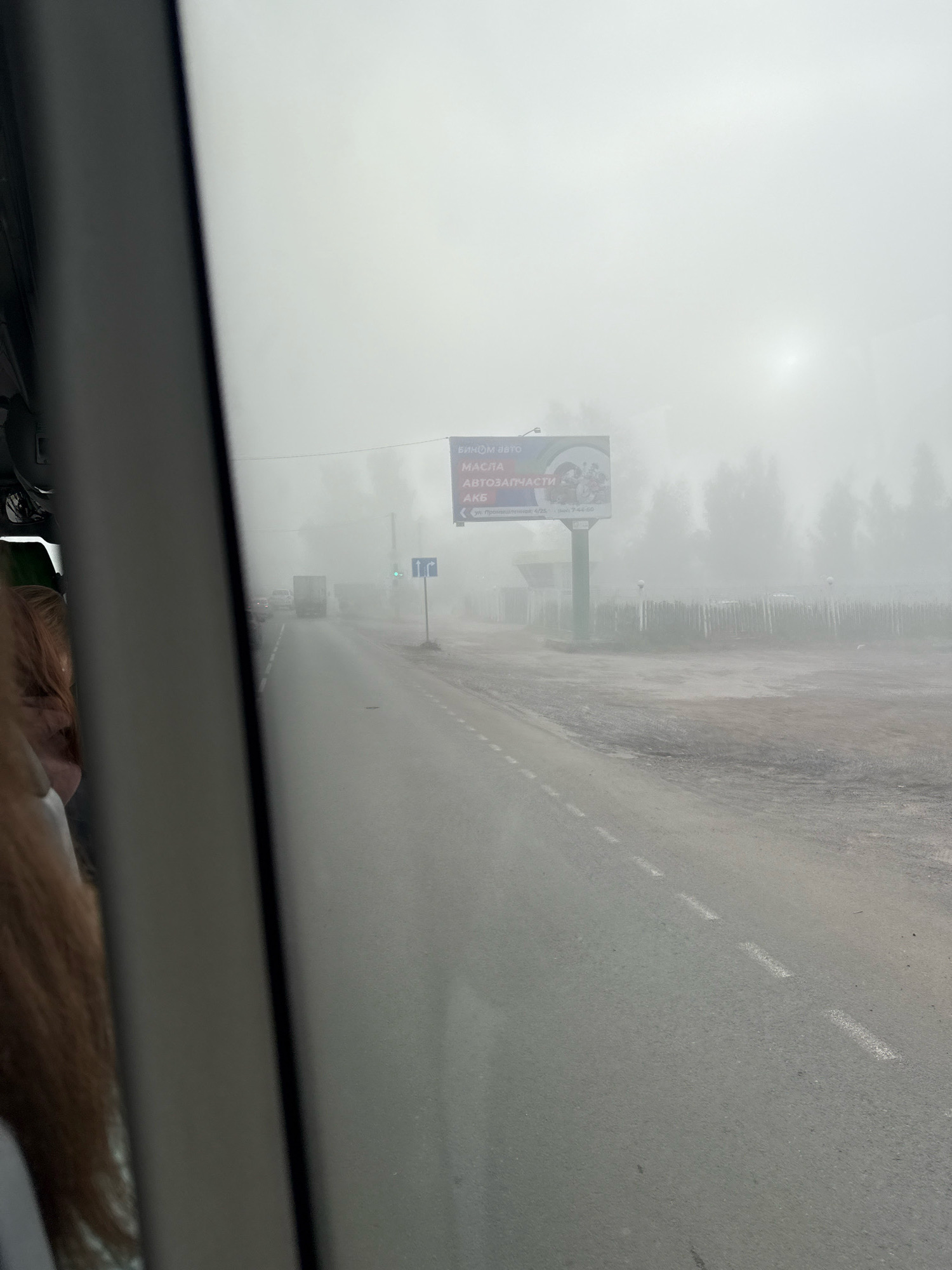 The image captures a foggy day on a road in Chaikovsky, Russia. A person's hair is visible on the left side of the frame, suggesting the photo was taken from inside a vehicle. The road is wet and appears to be a two-lane highway with white dashed lines. In the distance, several vehicles, including a truck, are on the road. A billboard advertising automotive parts and oils is visible on the right side of the road, partially obscured by the fog. A traffic sign with arrows indicating directions is also present. The environment is characterized by dense fog and trees lining the roadside.