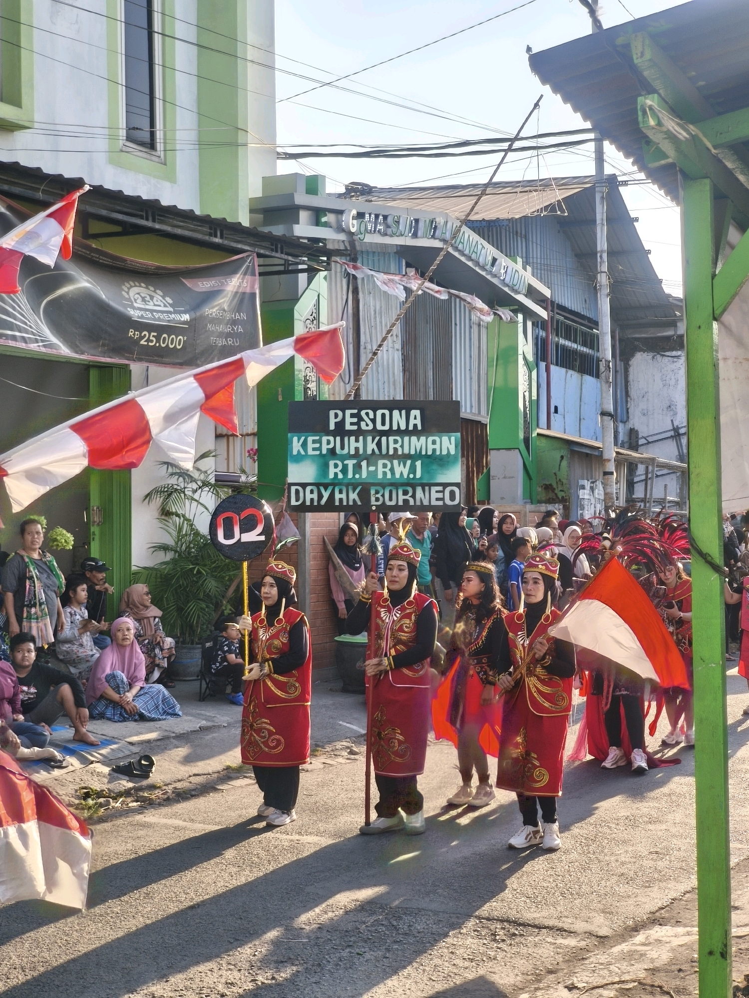 The image shows a street scene in Kepuh Kiriman, Indonesia, during what appears to be a parade or cultural event. Several people are present, including adults and children, many of whom are watching from the side of the street. In the foreground, three women in traditional red and gold attire are marching, holding flags and staffs. The woman on the left holds a round sign with the number "02" on it. Behind them, other participants in similar costumes can be seen, some with feathered headdresses. Flags, including Indonesian flags, are prominently displayed throughout the scene. Buildings with green and white facades line the street, and banners with text are visible. The lighting suggests it is daytime, with shadows cast on the asphalt road.