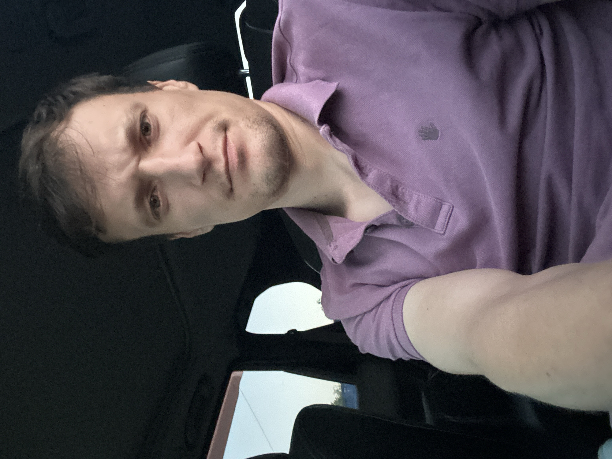 A man is sitting in the passenger seat of a car, looking at the camera. He is wearing a purple polo shirt. The car appears to be parked. Outside the car window, the sky is clear and bright, suggesting it is daytime. The interior of the car is dark, with the exception of the light coming through the window. The man's expression is neutral.