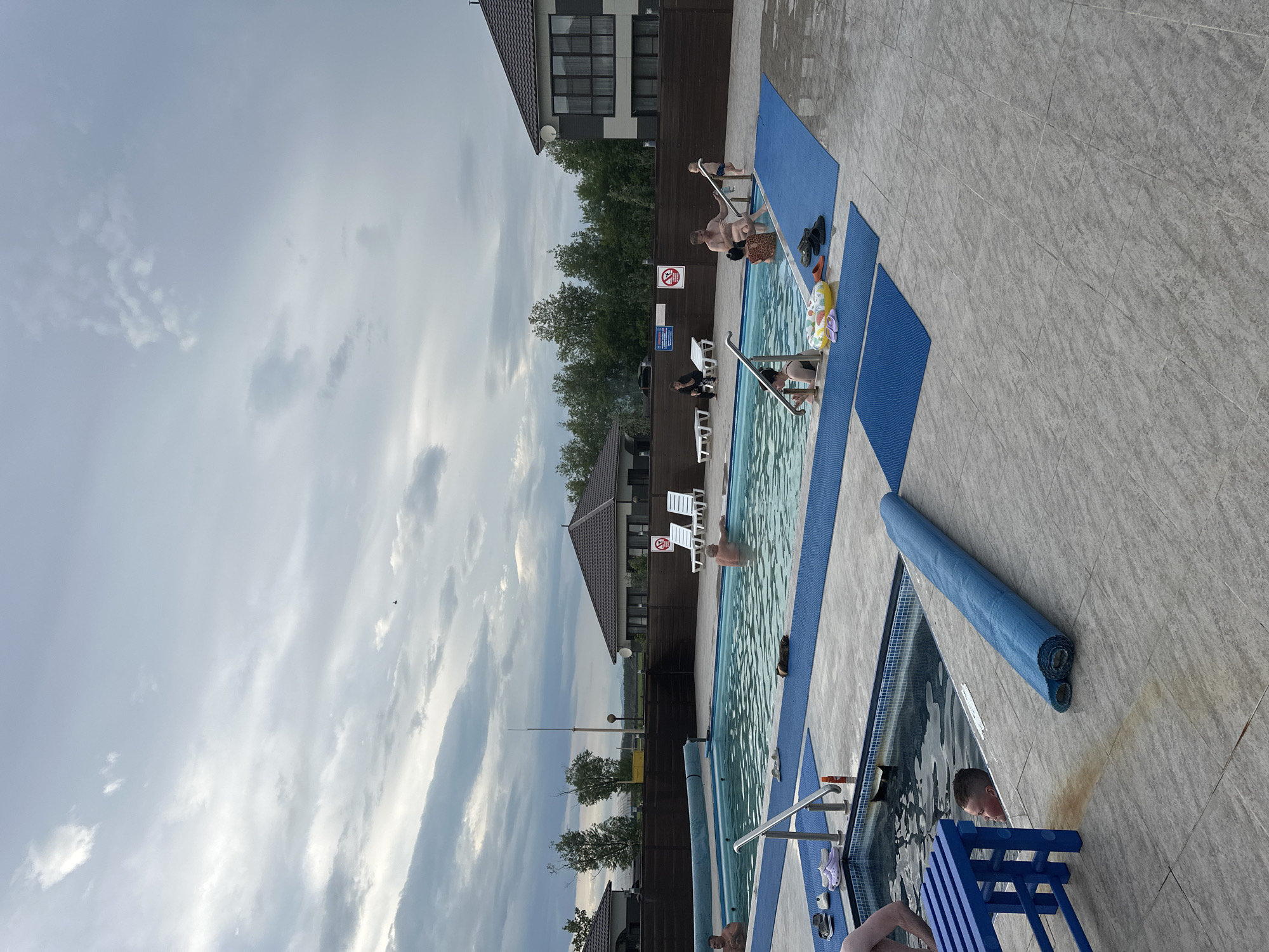 The image depicts an outdoor swimming pool area, likely in a resort or private residence setting. The pool is rectangular and filled with clear water. Several people are visible in and around the pool. In the main pool, two men are partially submerged, with one appearing to be standing and the other leaning against the side. A child is entering the pool from the steps, accompanied by an adult woman. Another woman is seated on the edge of the pool, interacting with a young child. Two lounge chairs are placed on the tiled area surrounding the pool, with two people seated on them. A blue bench is positioned near a smaller, square pool which also contains water and a child.
The pool area is paved with light-colored tiles, with blue mats laid out in sections. Objects such as flip-flops, a rolled-up blue mat, and a yellow floatation device are scattered on the ground. In the background, there are modern houses with dark roofs and visible windows, surrounded by a wooden fence and trees. The sky is overcast with dramatic cloud formations. The scene suggests a relaxed, recreational atmosphere, possibly during a summer day, given the presence of swimwear and swimming activities.