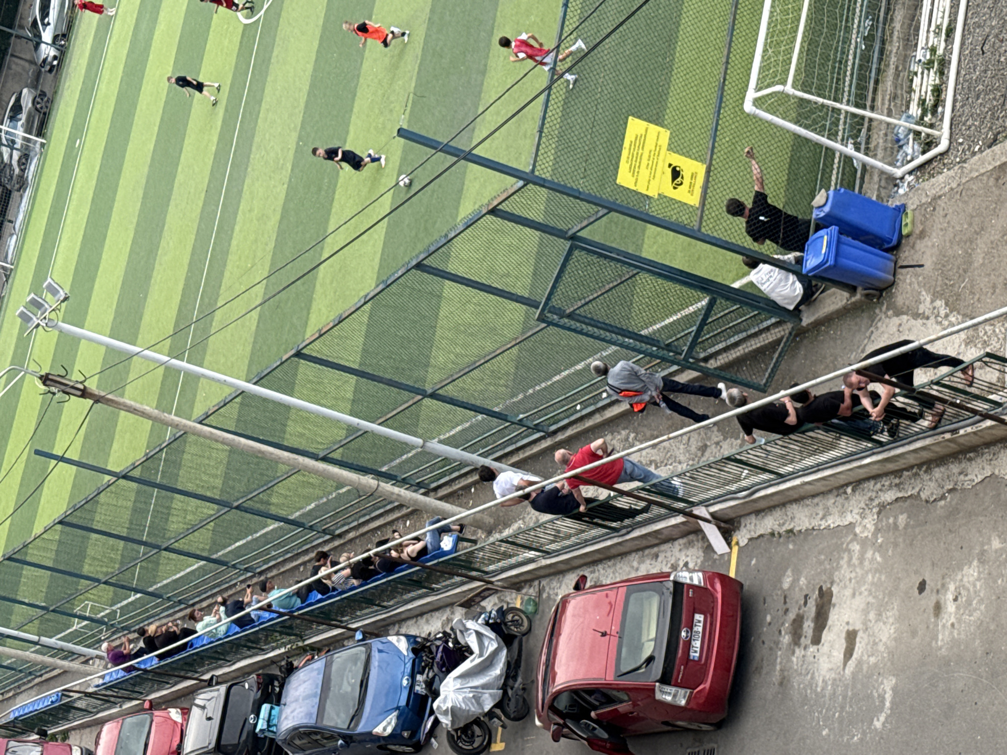 This image depicts an outdoor soccer field in Tbilisi, Georgia. A group of people are observing a soccer game from beyond a fence. Some individuals are seated on bleachers, while others stand along the fence. On the field, several players are engaged in a match, some in black uniforms and others in red and white. Adjacent to the soccer field, there is a parking area with several cars, including a red Toyota Prius, a blue hatchback, and a black sedan. A motorcycle covered with a silver tarp is parked next to the blue car. Blue recycling bins are placed near the fence, close to a soccer goal. The ground surface appears to be a mix of concrete and asphalt. A yellow sign with text and an illustration is affixed to the fence. The overall scene captures a casual sporting event with spectators and vehicles present.