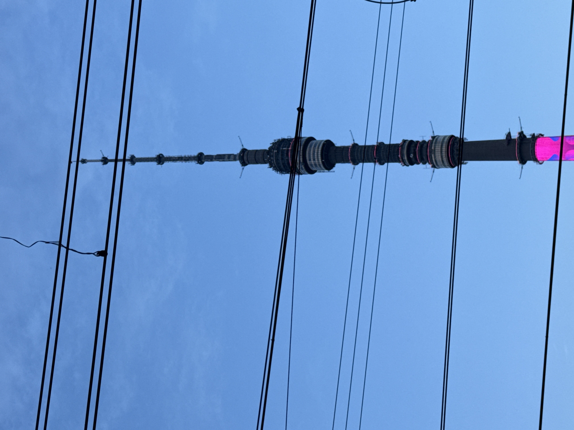 The image depicts the Ostankino Tower in Moscow, Russia, viewed from a low angle looking up. Power lines run across the foreground of the image, partially obscuring the tower. The tower itself is a tall, thin structure with multiple cylindrical sections along its length. The top of the tower tapers to a point. One of the sections of the tower is illuminated with pink lights. The sky is a light blue color.