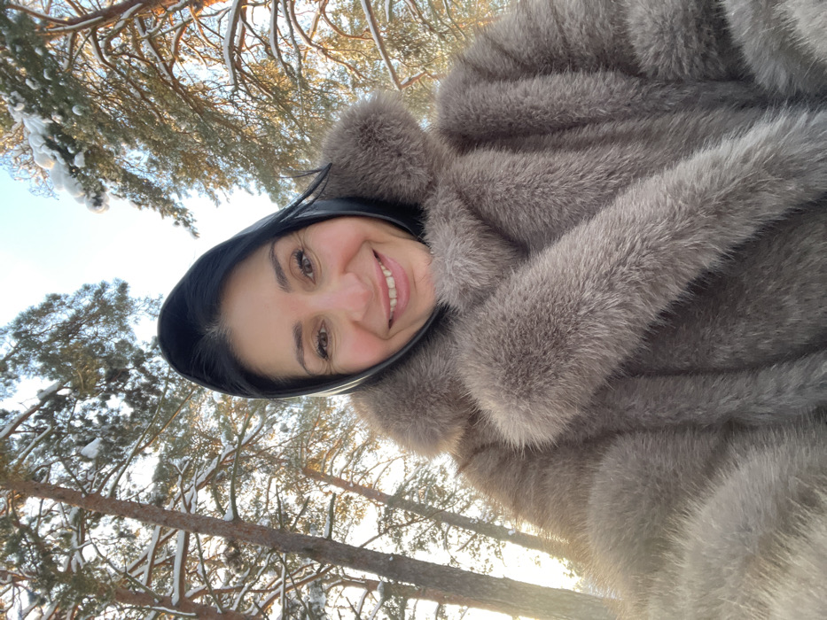 The image presents a close-up selfie of an adult female in a snowy forest environment in Syasstroy, Russia. The subject, viewed from the upper chest upwards, has dark hair partially covered by a black head covering. She is wearing a thick, light brown/grey fur coat featuring a large collar. Her facial expression displays a broad smile, with her eyes directed toward the camera. The background consists of numerous tall evergreen trees, their branches visibly laden with snow, under a bright, clear sky. Sunlight emanates from the left side of the frame, illuminating portions of the snow-covered foliage.