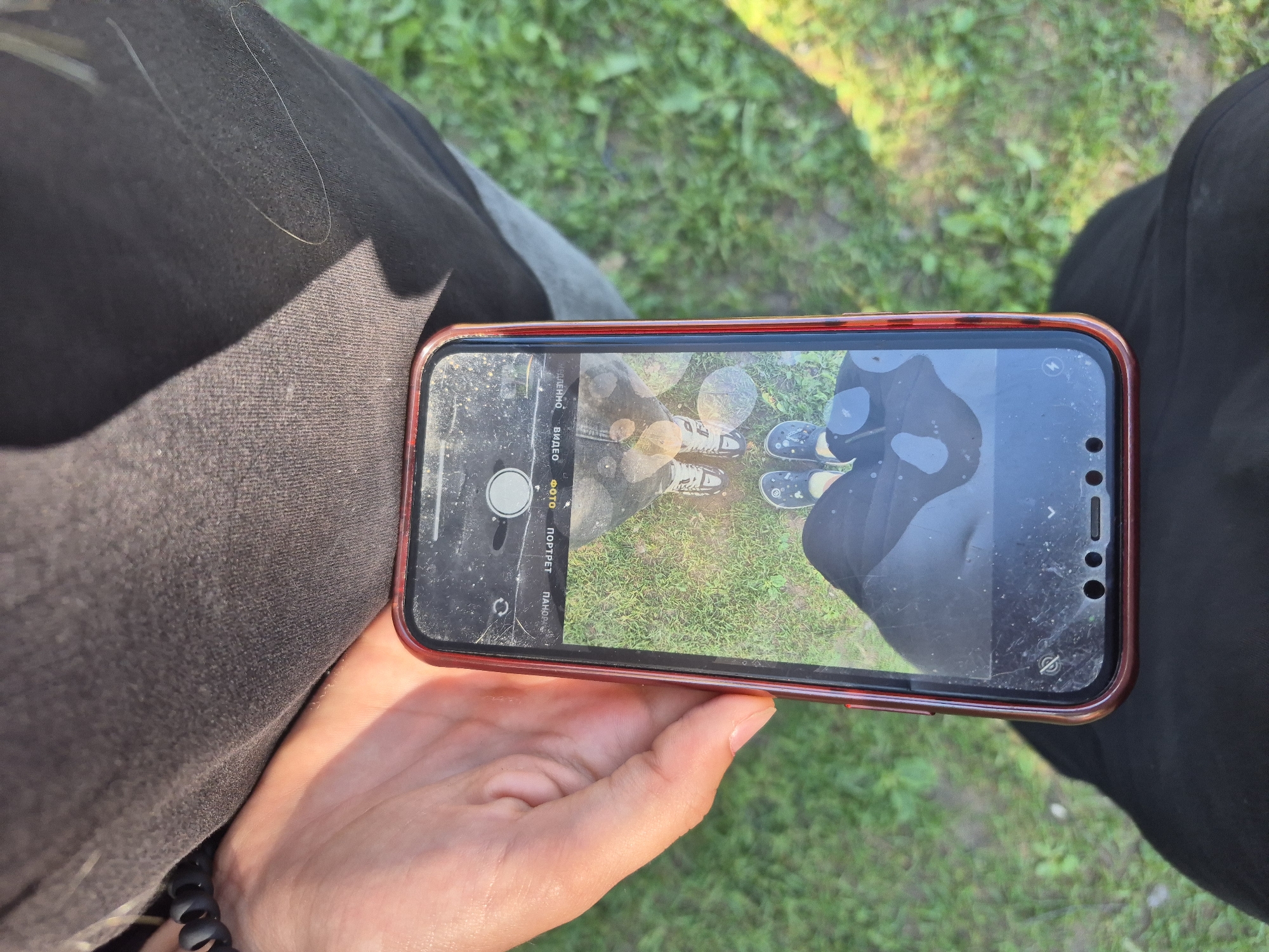 A person holds a smartphone with a cracked screen, displaying a selfie of their feet and the ground. The phone is in portrait mode, and the camera interface is visible, showing options for "PHOTO," "PORTRAIT," and "VIDEO." The background is green grass. The image is taken outdoors during the daytime. The person is wearing dark pants and dark shoes. The phone has a red casing. There is no visible indication of the specific location within Chaikovsky, Russia.